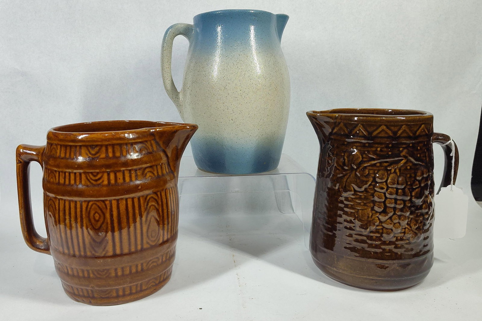 3 American Stoneware/Brownware Pitchers: This lot consists of a trio of ceramic pitchers featuring various glaze techniques and relief-molded decorations, likely of 20th-century American manufacture. The first item is a stoneware pitcher wit