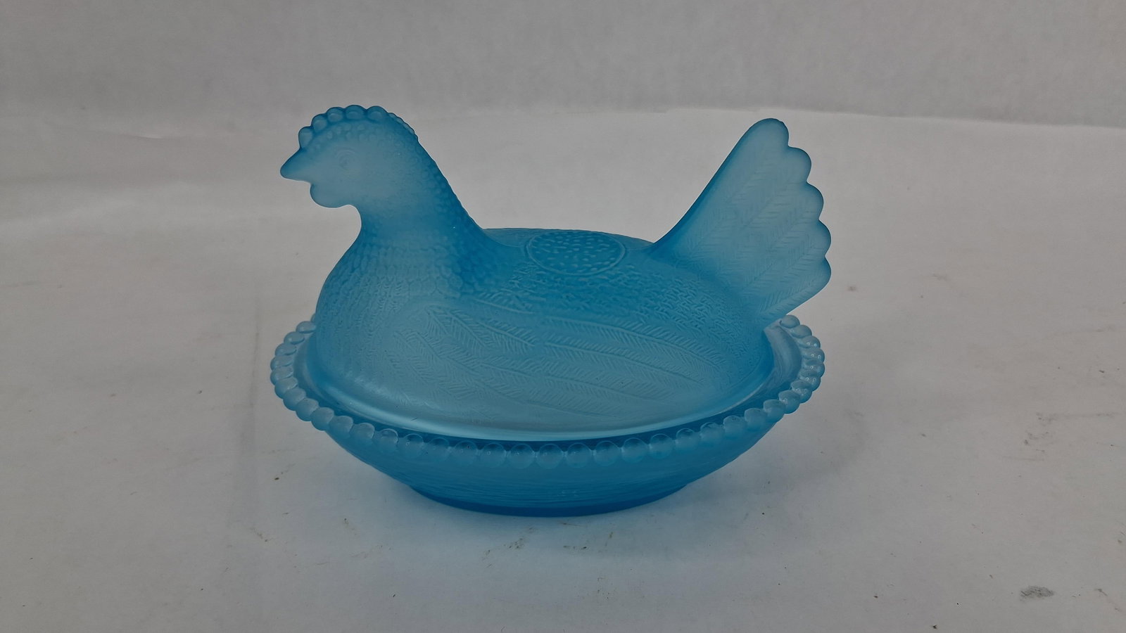Blue Satin Glass Hen on Nest Dish: 4.5 in long A two-piece Hen on Nest covered dish crafted from medium blue pressed glass with a satin or frosted finish. The lid is molded in the form of a seated hen featuring detailed plumage on the
