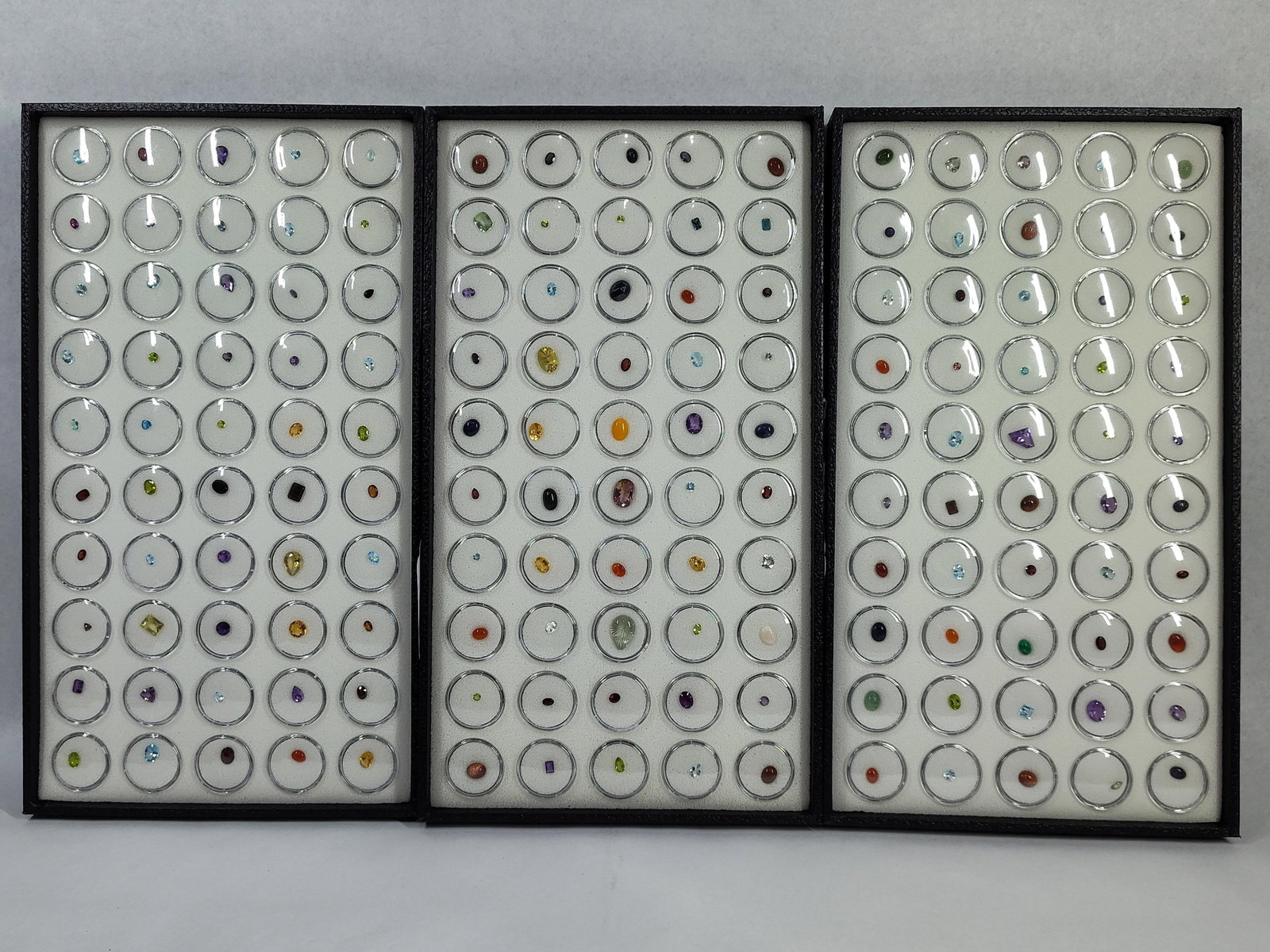 150 Faceted & Cabochon Gemstones, Trays: An extensive collection of approximately 150 loose gemstones organized into three matching black professional display trays. Each tray features a 10 by 5 grid of clear plastic gem jars with white