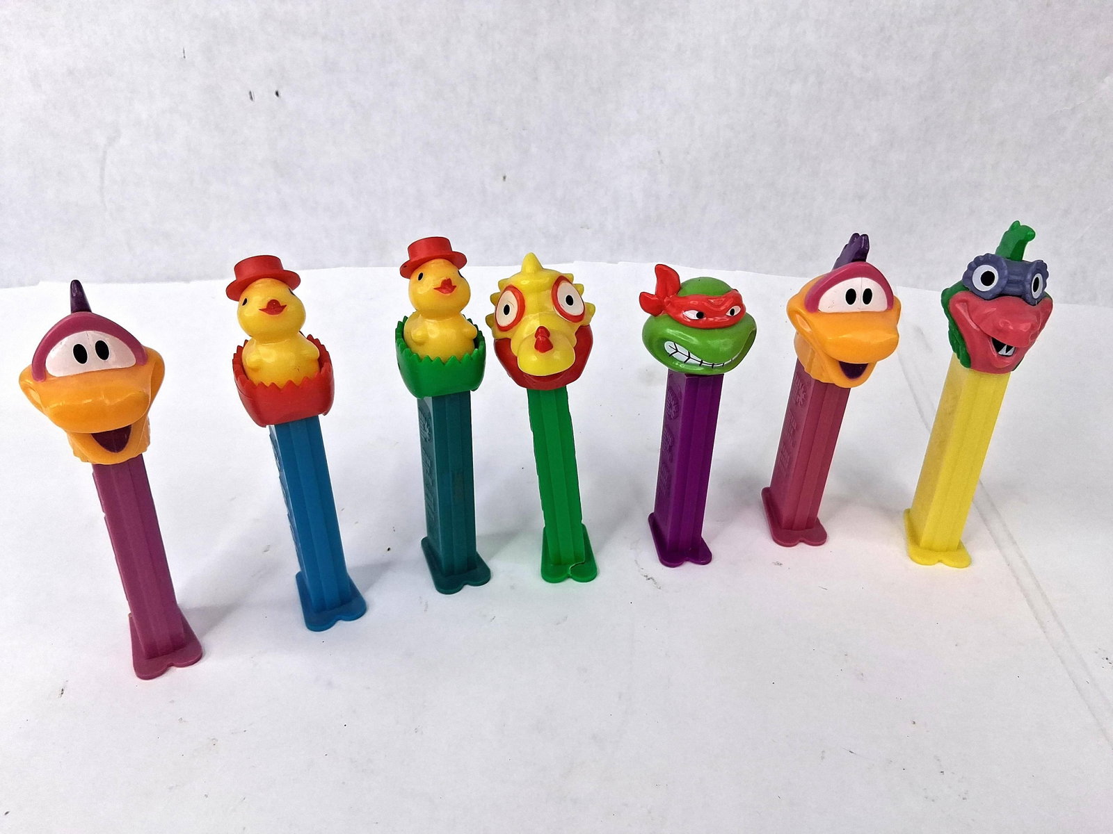 7 PEZ Dispensers, TMNT & Pez-A-Saurs: A collection of seven loose PEZ candy dispensers constructed from molded plastic with spring-loaded tilt-head mechanisms. The assortment includes a licensed Teenage Mutant Ninja Turtles character,