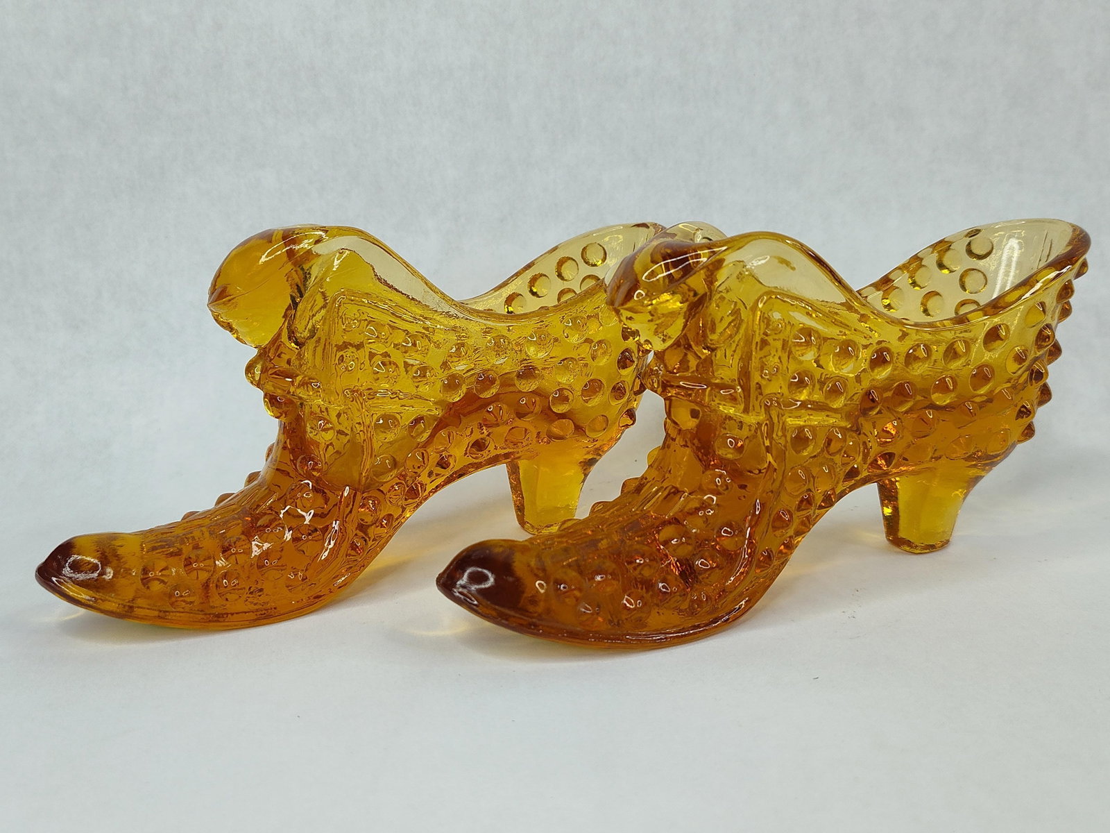 Pair Fenton Amber Hobnail Cat Slippers: A pair of Fenton Art Glass decorative shoe slippers in transparent amber. These items are crafted from pressed glass featuring an all-over hobnail texture. The form is modeled after a Victorian