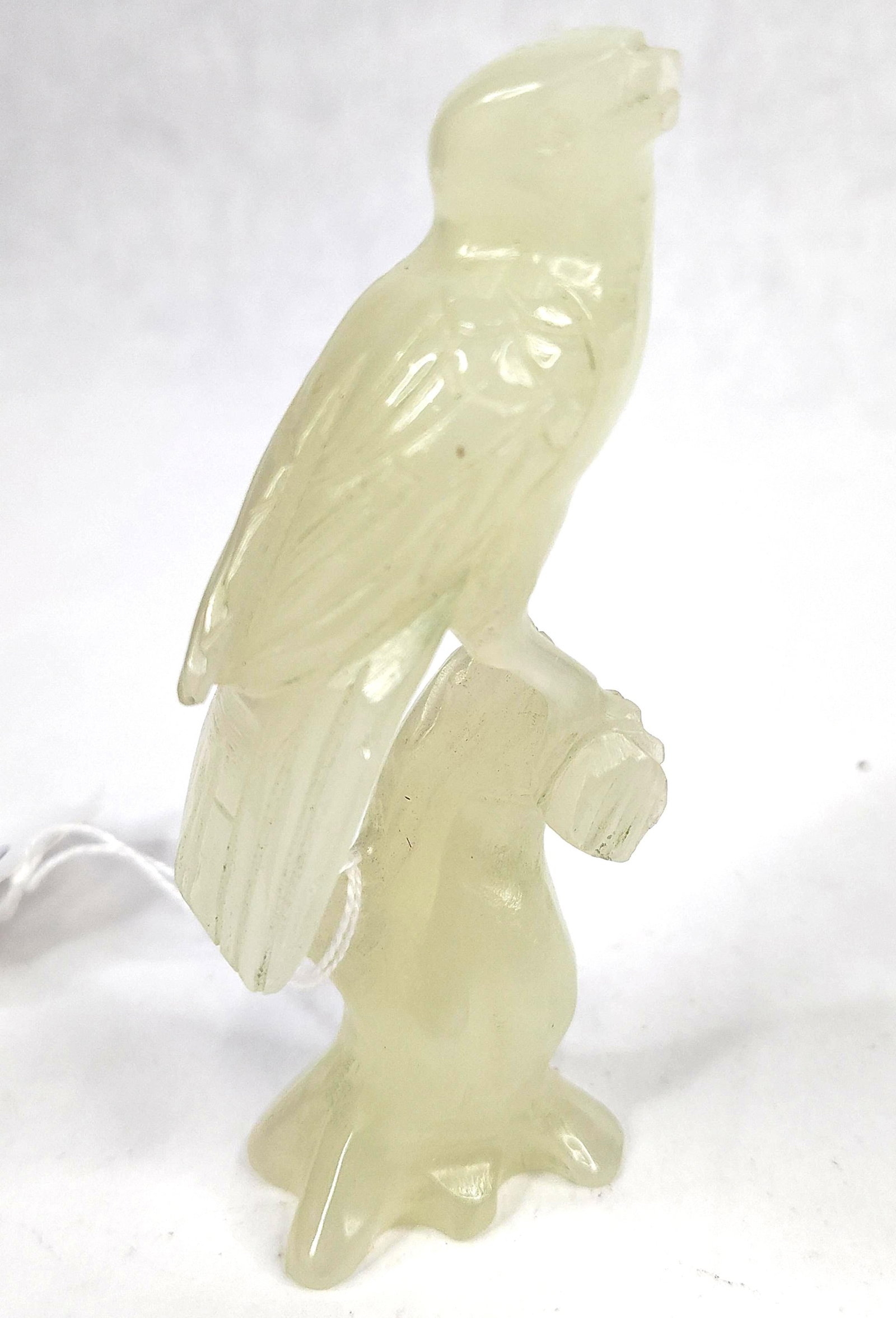 Chinese Pale Celadon Jade Bird of Prey: This hand-carved stone figurine depicts a bird of prey, likely a falcon, perched upon a textured branch. The piece is fashioned from a translucent pale celadon green stone, consistent in appearance wi