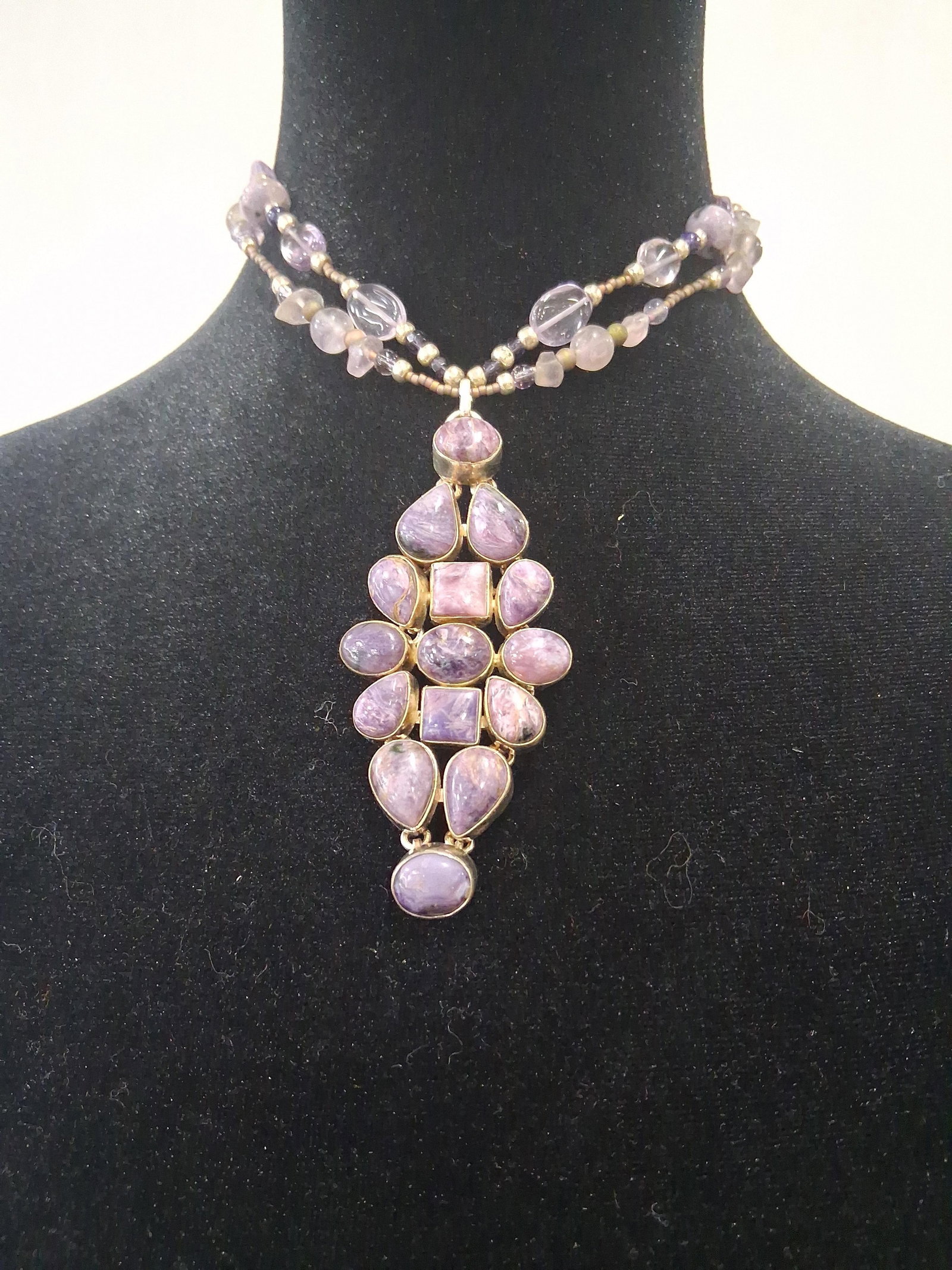 Str. Charoite & Amethyst Multi Necklace: This jewelry item is a multi-strand beaded necklace featuring a large, articulated sterling silver pendant. The pendant contains thirteen bezel-set cabochon gemstones of various shapes, including