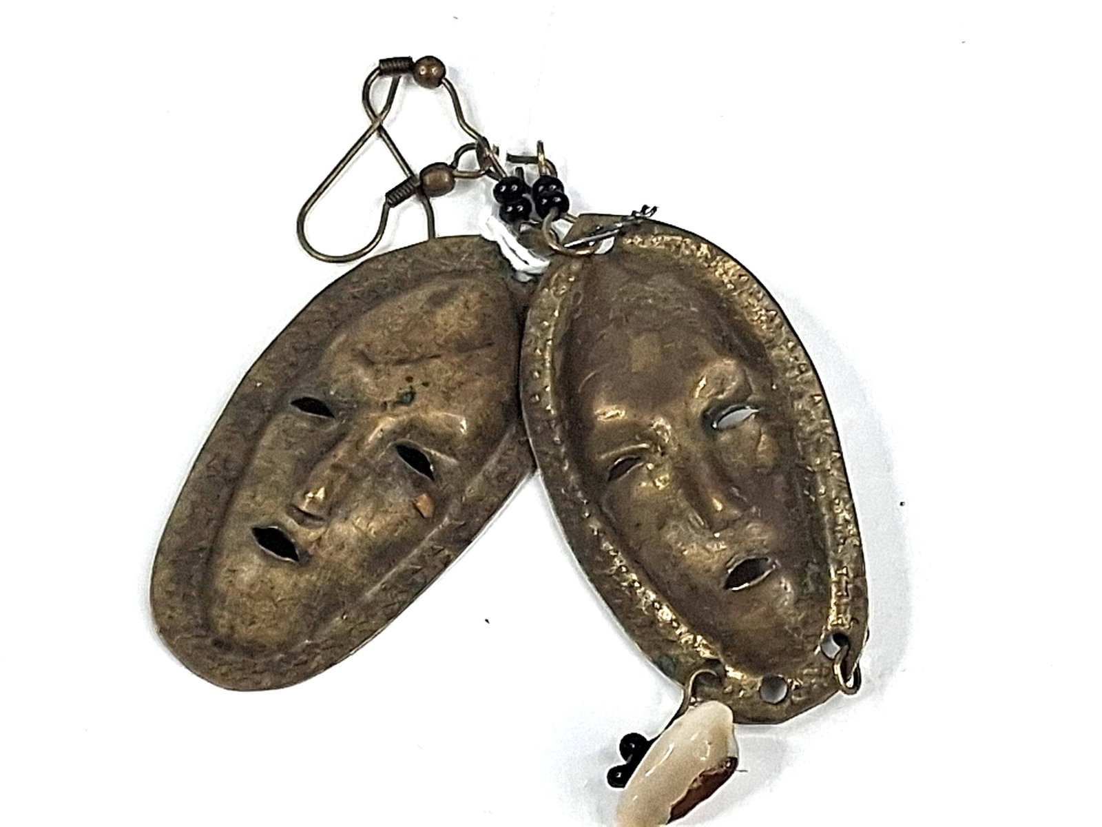Pair Mask Drop Earrings w/ Cowrie Shell: A pair of drop earrings featuring stylized mask pendants crafted from a brass-toned metal. Each mask is oval in shape, featuring a repousse-style elongated face with slit-cut eyes and an open mouth.