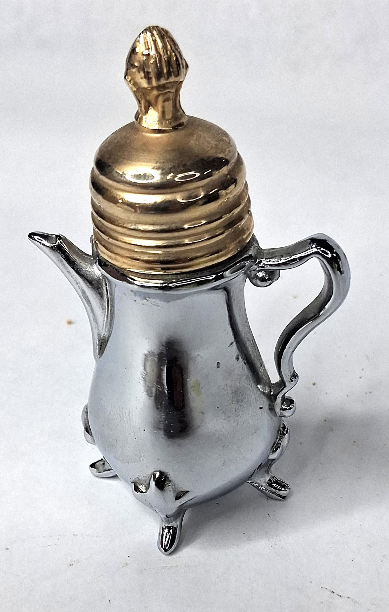 Kutmaster by Cory Chrome Teapot Lighter: A miniature novelty table lighter in the form of a teapot, produced by Kutmaster by Cory. The item features a chromium plated base metal body with a footed base, a scrolled handle, and a short spout.