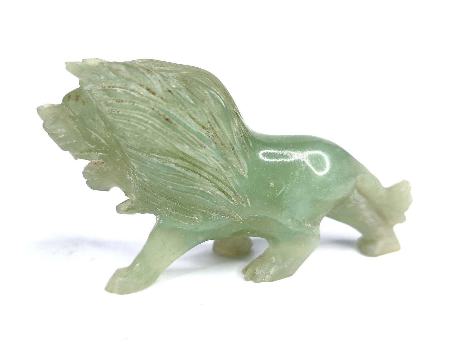 Carved Green Hardstone Lion Figurine: A carved hardstone figurine depicting a male lion in a dynamic, low-slung striding pose. The piece is fashioned from a translucent, pale celadon-green stone featuring a polished, waxy luster. The carv