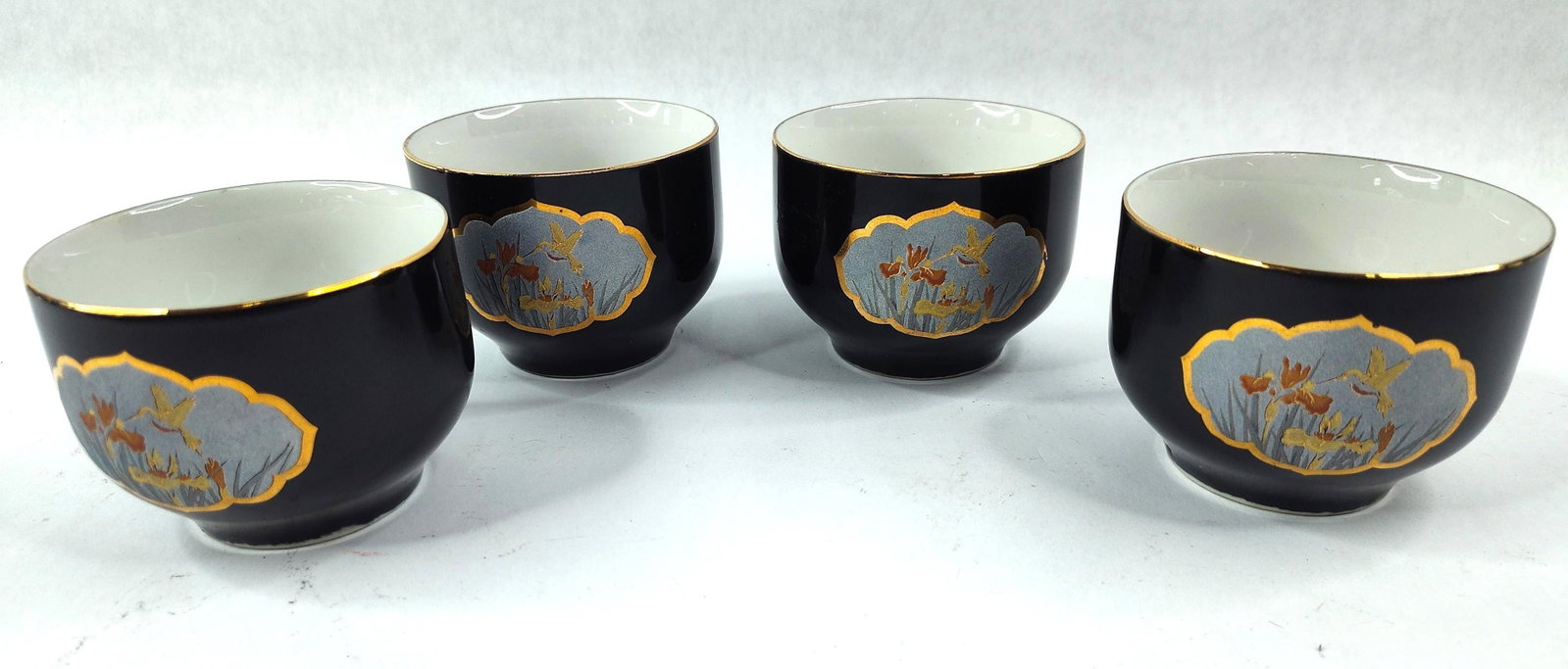 4 Kutani Porc. Hummingbird Sake Cups: A set of four porcelain sake cups, or ochoko, manufactured for the export market during the mid-20th century. Each cup features a deep black-glazed exterior and a white-glazed interior with a gilded