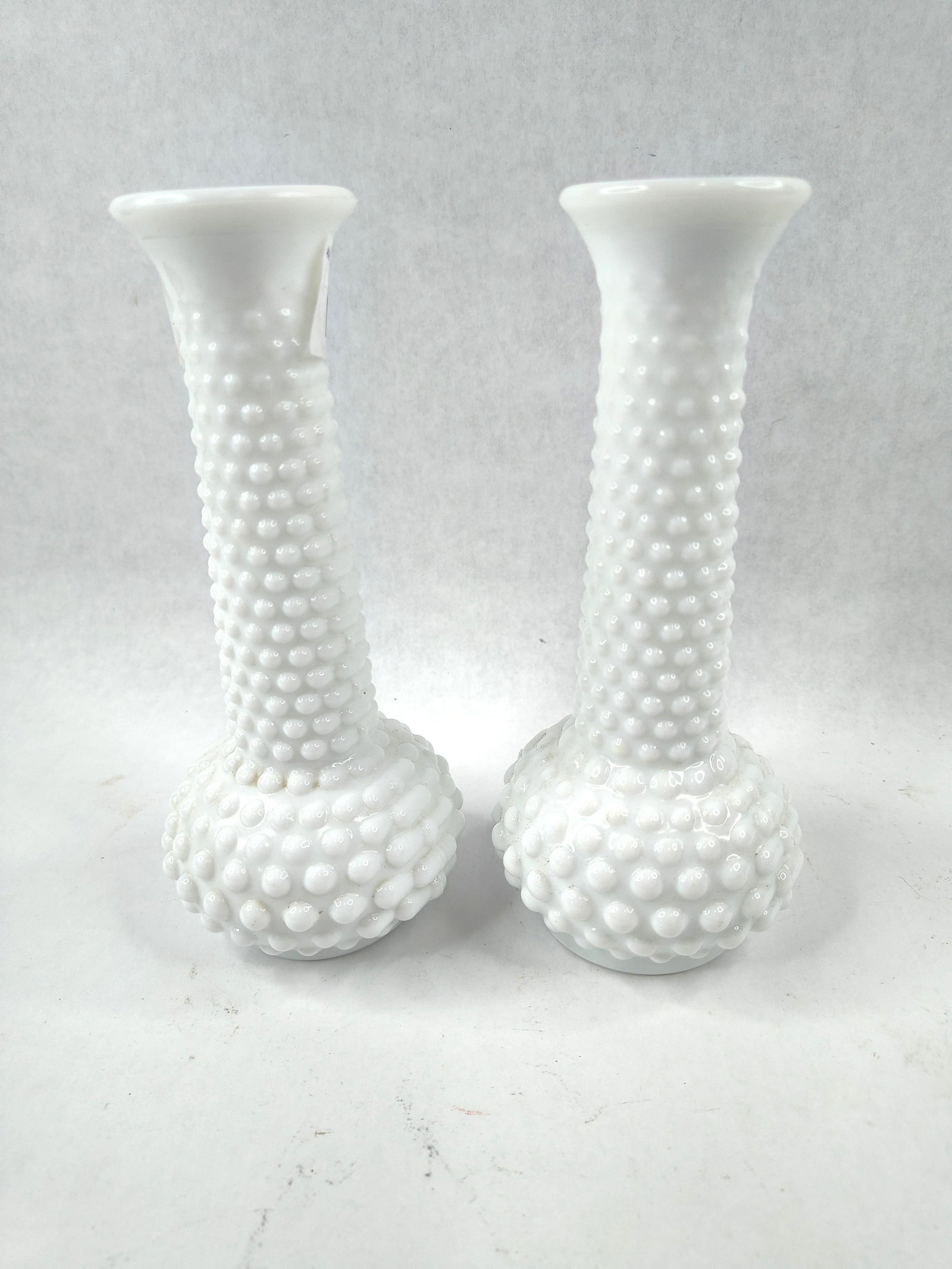 E.O. Brody Milk Glass Hobnail Bud Vases: This lot features a pair of white milk glass bud vases manufactured by the E.O. Brody Company. The vases exhibit a classic hobnail pattern across a bulbous base and an elongated, slender neck that fin