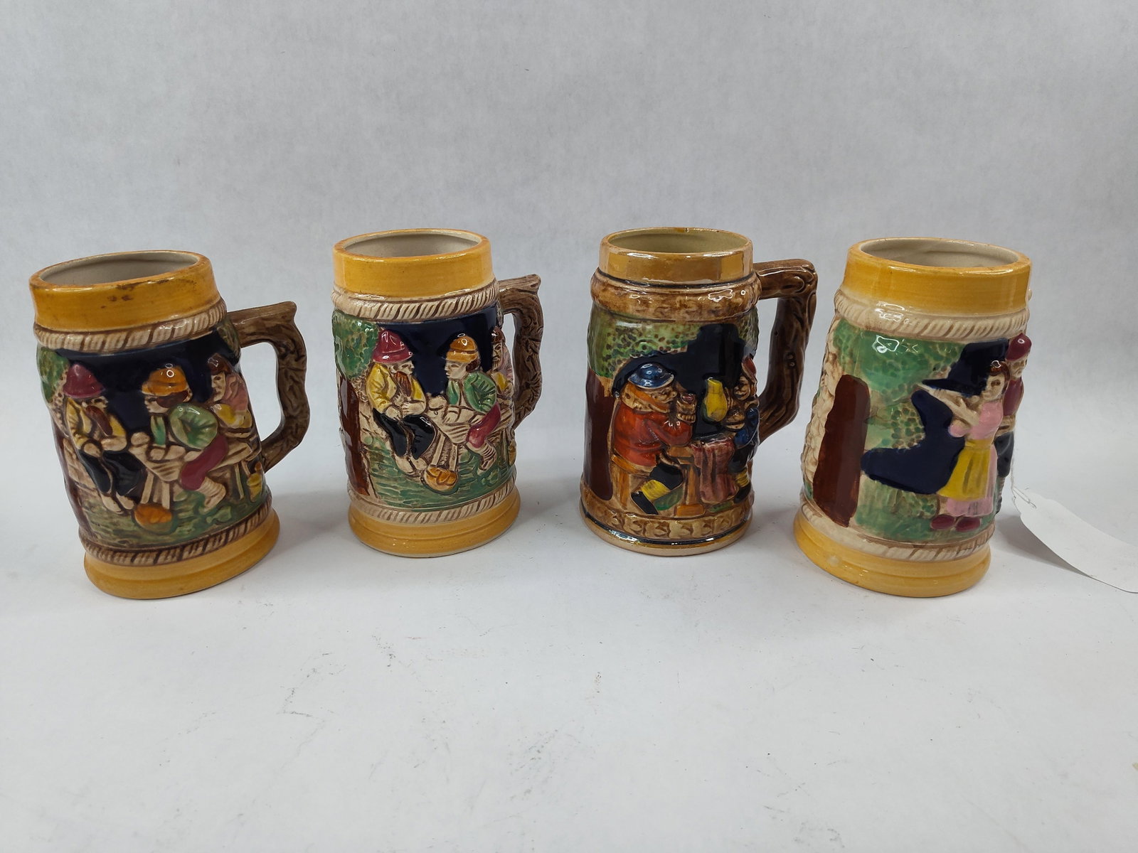 Set 4 Fairway Japanese Ceramic Steins: This set of four miniature glazed earthenware beer steins features relief-molded decoration in a German souvenir style. Each vessel is designed in a tapered cylindrical form with a molded handle