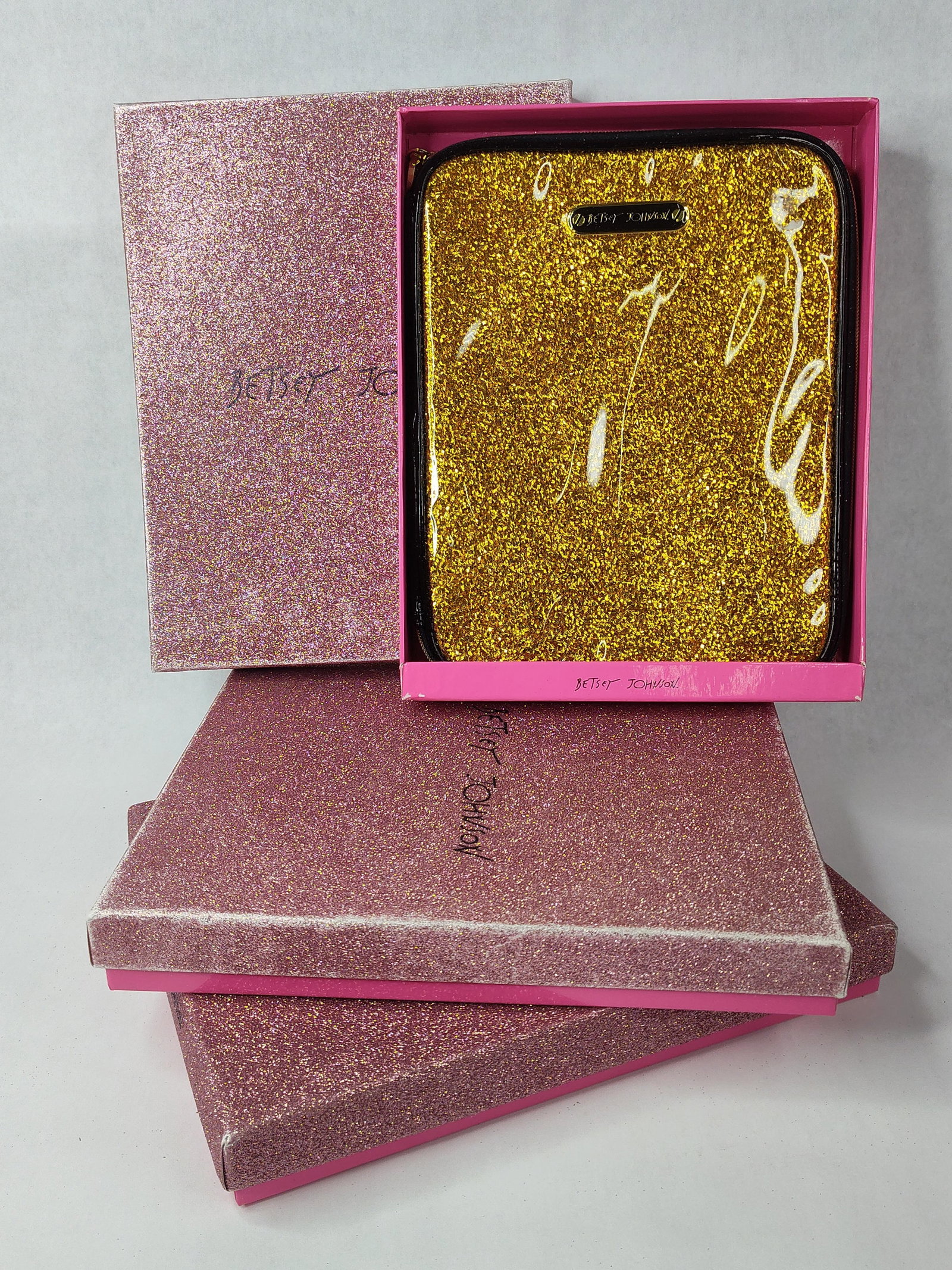 Betsey Johnson Gold Glitter iPad Case: A gold-tone glitter iPad folio case by Betsey Johnson. The exterior is constructed of synthetic gold glitter encased in clear vinyl with a black textile zipper tape and gold-tone metal hardware. A
