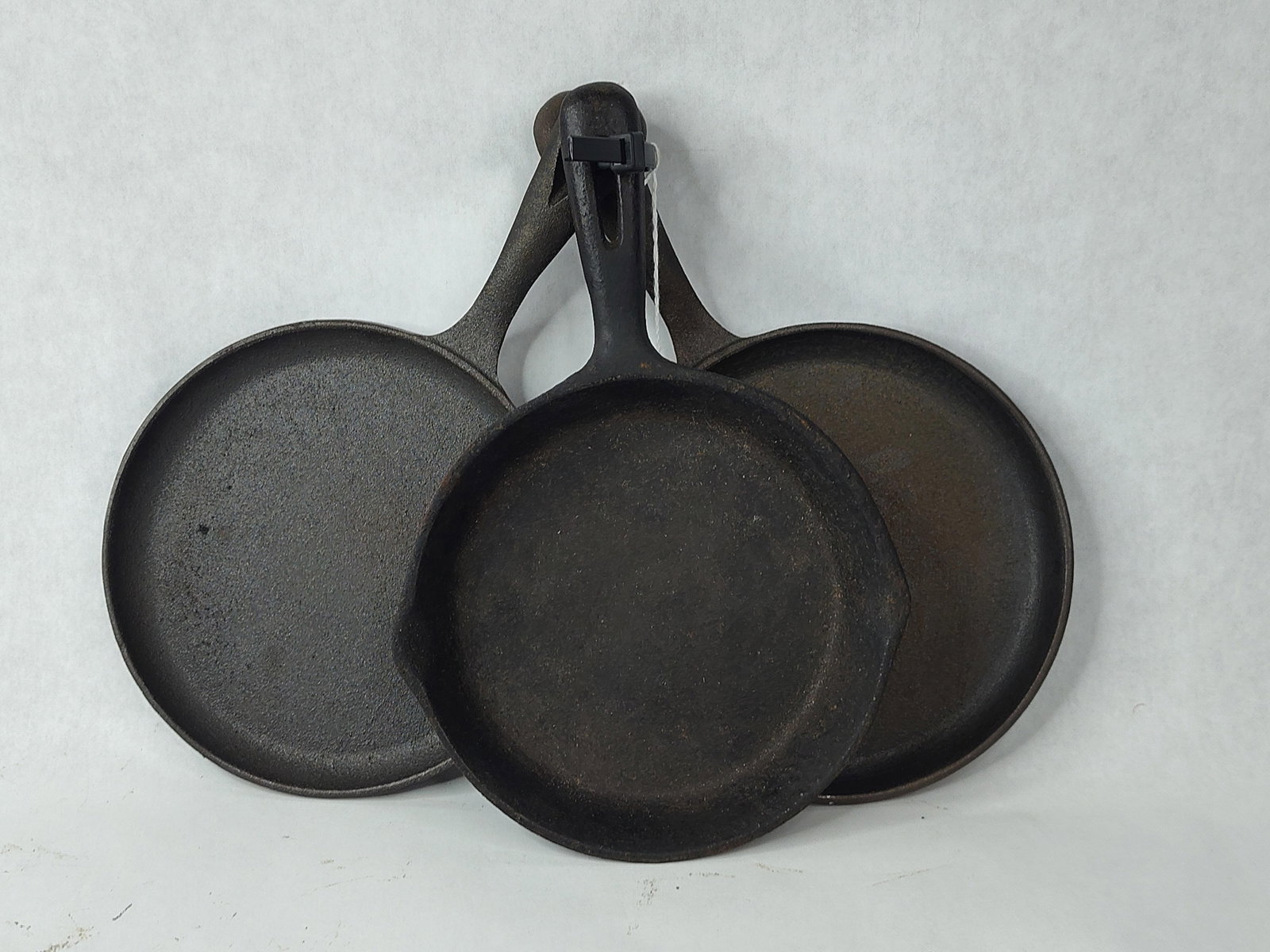 Trio Vtg Cast Iron Griddles & Skillet: A collection of three cast iron cookware items, consisting of two circular griddles and one skillet. The griddles are characterized by shallow rims and integral handles with elongated hanging slots.