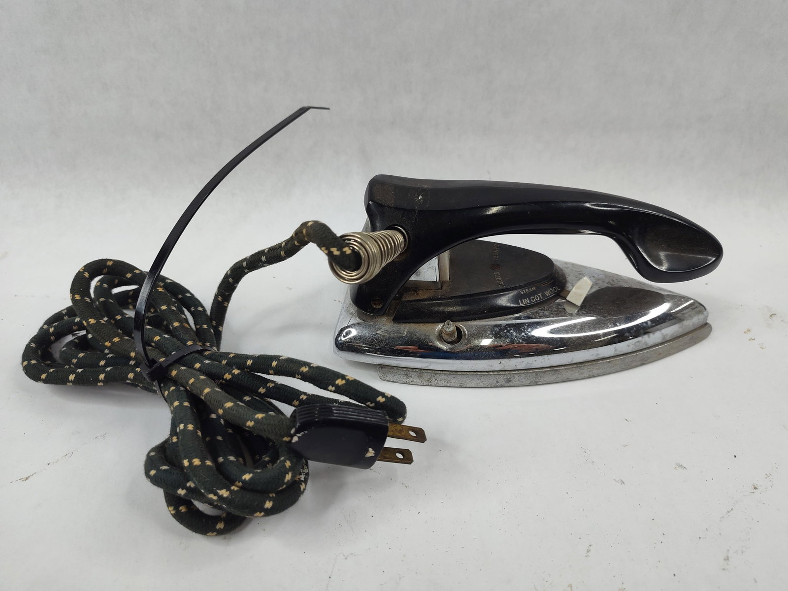 General Electric 11F37 Steam & Dry Iron: A General Electric electric steam and dry iron, catalog number 11F37. The unit features a chrome-plated metal shell with a black thermoset plastic handle. It retains its original cloth-wrapped power c