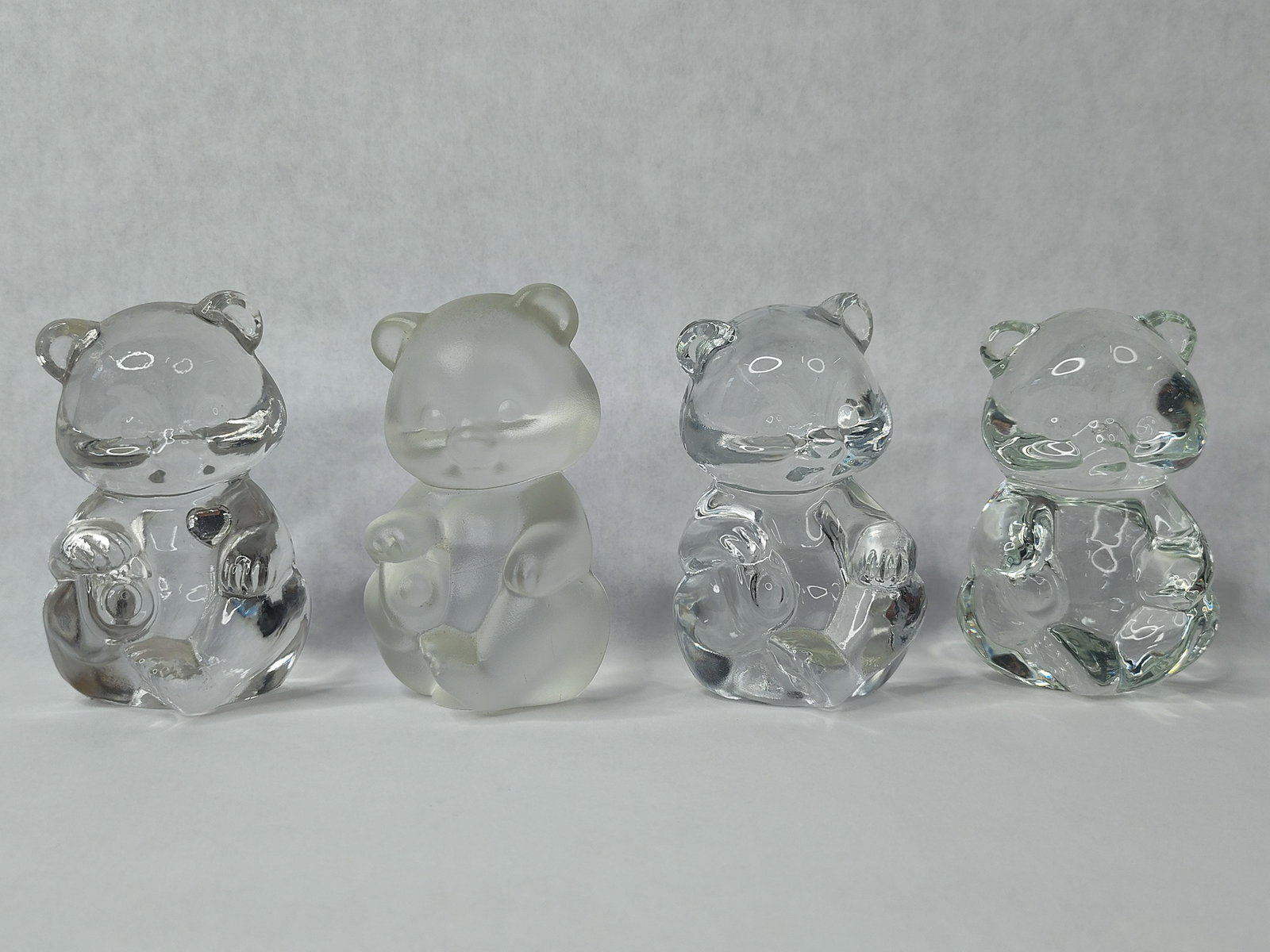 Set 4 Fenton Art Glass Seated Bear Figs: This collection consists of four seated bear figurines manufactured by the Fenton Art Glass Company. The set includes three figurines crafted in clear flint glass and one figurine in a frosted satin g
