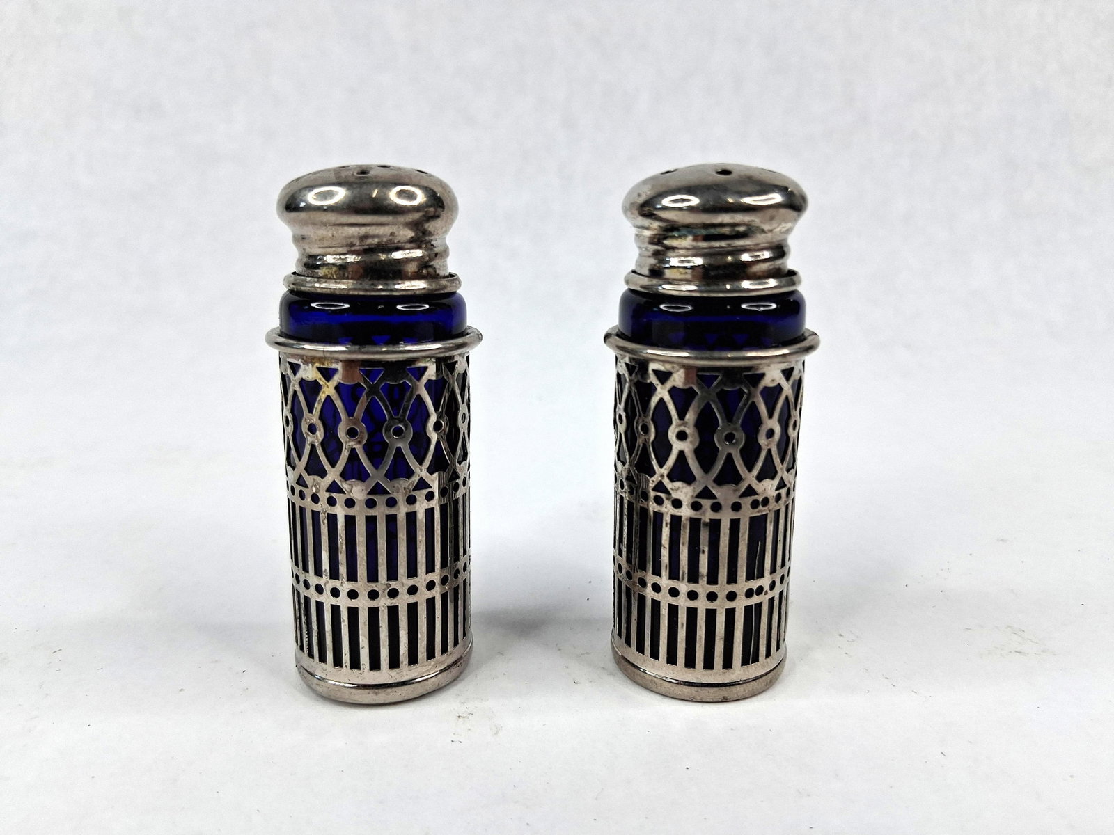 Pair Cobalt Glass & Silver-Tone Shakers: This lot consists of a matching pair of salt and pepper shakers, each featuring a cylindrical cobalt blue glass liner encased in a reticulated metal sleeve. The sleeves are decorated with a two-part p