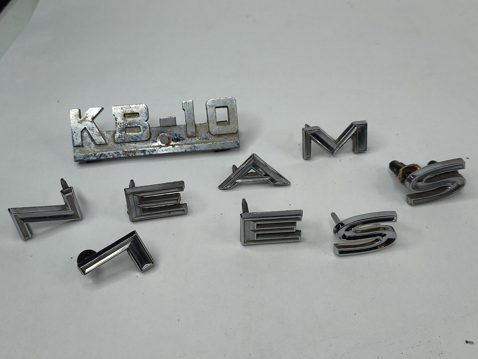 9 Vtg Chrome Auto Emblems & KB-10 Badge: This collection consists of nine chrome-plated die-cast metal automotive emblems and letters. The primary piece is a fender model designation badge for an International Harvester KB-10 heavy-duty truc