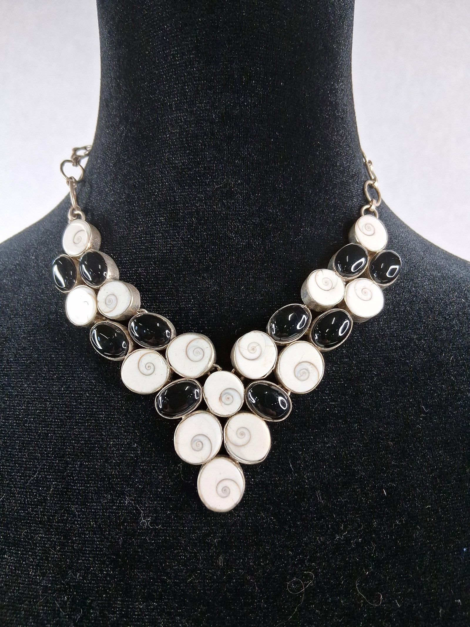 Shiva Eye Shell & Onyx Bib Necklace: A tiered V-shaped bib necklace composed of twenty-two linked bezel-set units. The design features fourteen oval Shiva eye (operculum) shells displaying their natural spiral patterns alongside eight