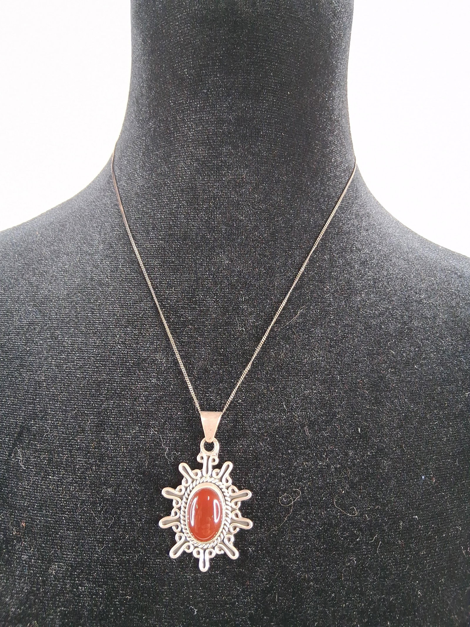 model: This sterling silver necklace features a pendant with a sunburst motif and an Italian curb link chain. The pendant centers an oval cabochon-cut carnelian gemstone, bezel-set within a twisted wire bord