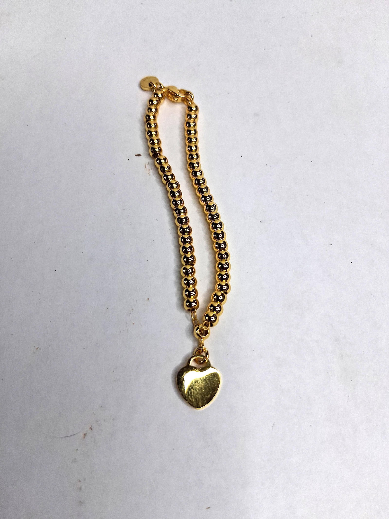 Gold-Toned Bead Bracelet w/ Heart Charm: A yellow gold-toned bead bracelet composed of a row of uniform spherical beads on a flexible internal structure, terminating in a lobster claw clasp. The bracelet features a fixed heart-shaped charm a