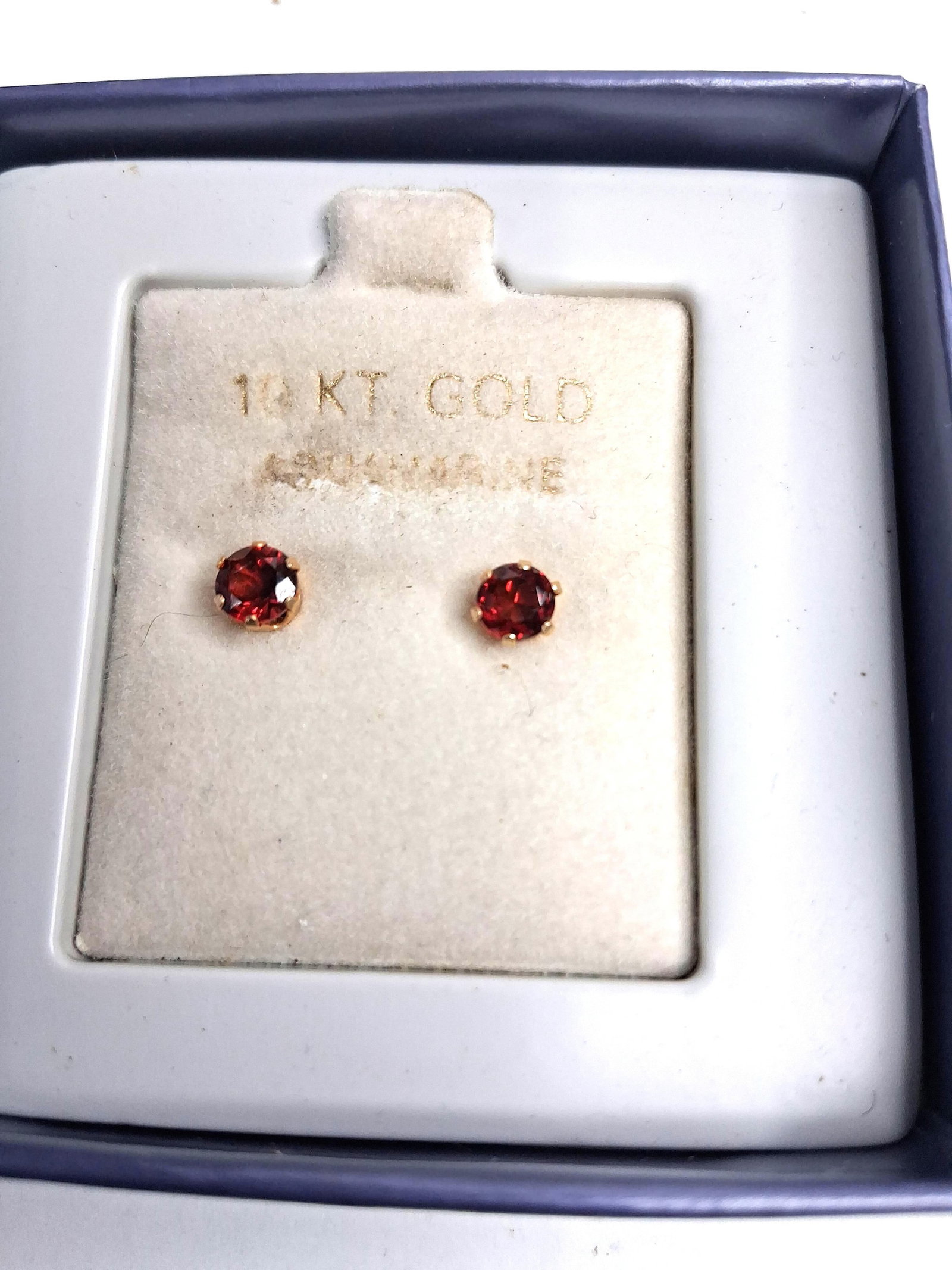 Pair 14kt Yellow Gold & Garnet Studs: A matched pair of gemstone stud earrings, each featuring a round-cut, dark red garnet. The stones are secured in six-prong basket settings of 14kt yellow gold. The earrings are mounted on a cream-colo