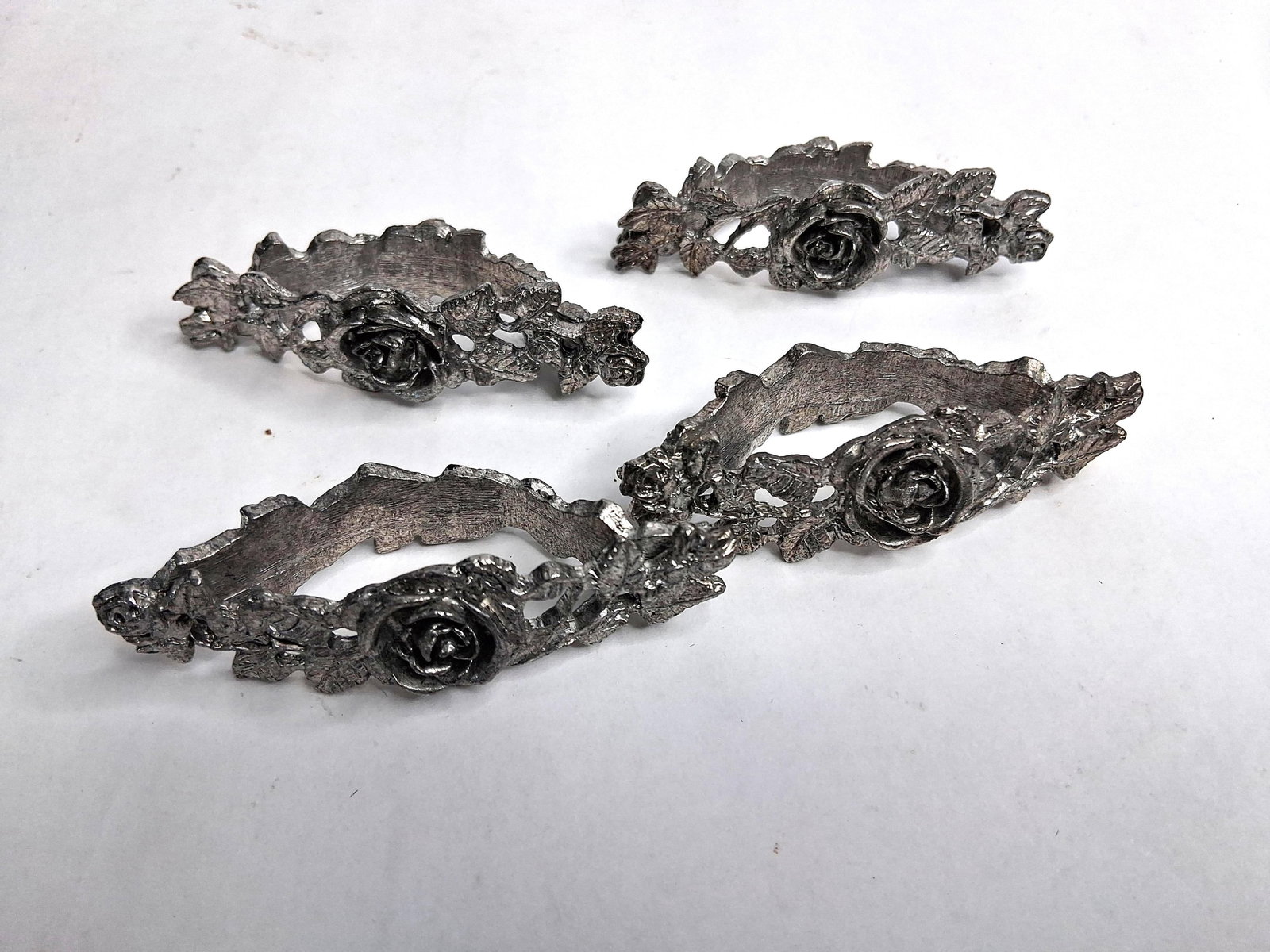 Metzke Inc. Pewter Rose Napkin Rings: A set of handcrafted cast pewter napkin rings by Metzke, Inc. Each ring features a prominent central rose blossom flanked by detailed foliate and vine motifs with a rough-hewn, textured finish. The in