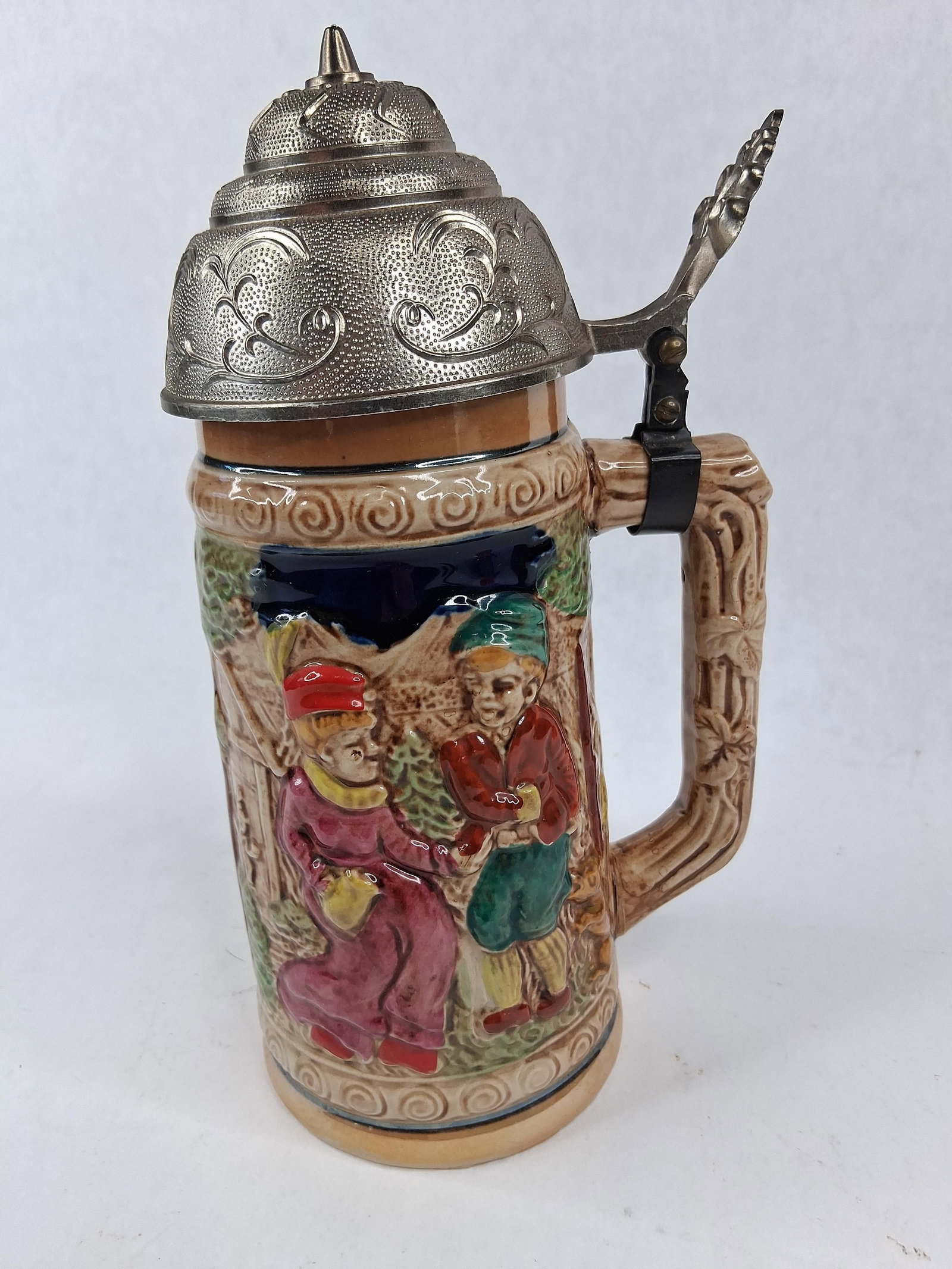 Vtg Musical Ceramic Alpine Relief Stein: This musical lidded beer stein features a relief-molded and hand-painted glazed earthenware body. The central decoration depicts a traditional Bavarian or Alpine scene with a seated woman in a maroon 