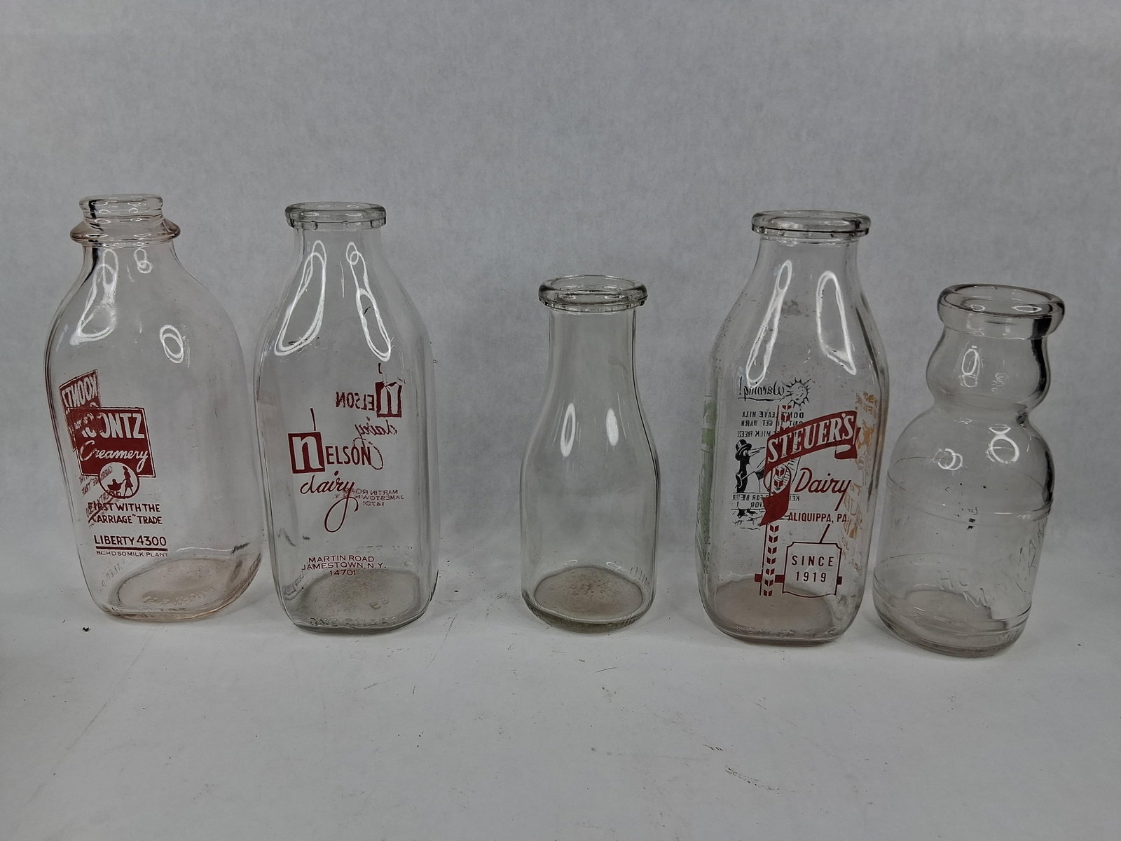 Regional American Glass Milk Bottles: This collection consists of clear glass milk bottles from various regional United States dairies, showcasing both square and round forms along with applied color labels and embossed details. The lot i