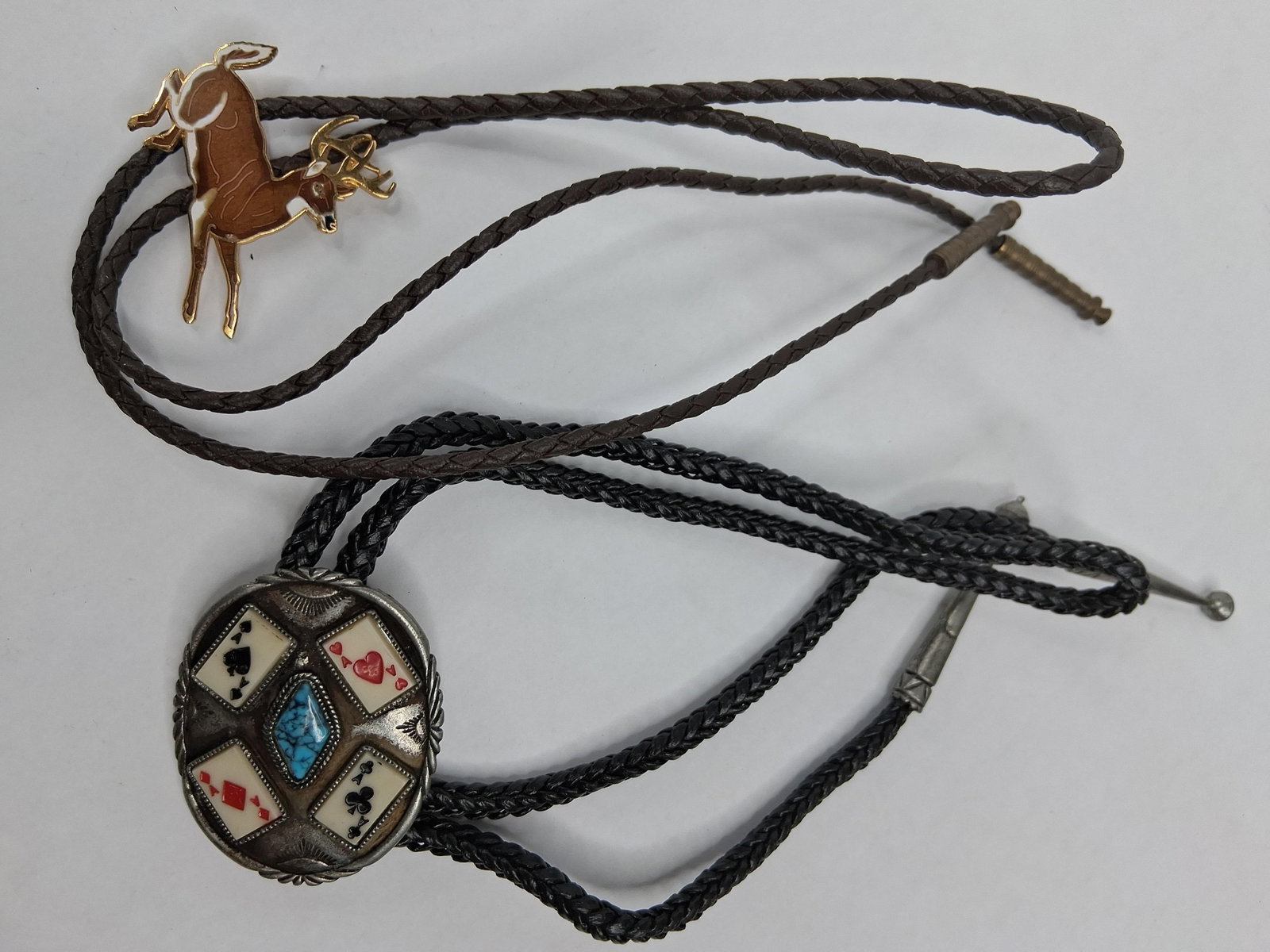 Pair Vtg Western Bolo Ties: Buck & Aces: This lot consists of two Western-style bolo ties. The first bolo tie features a gold-tone base metal slide designed as a jumping buck decorated with brown and white enamel. The slide is attached to a 