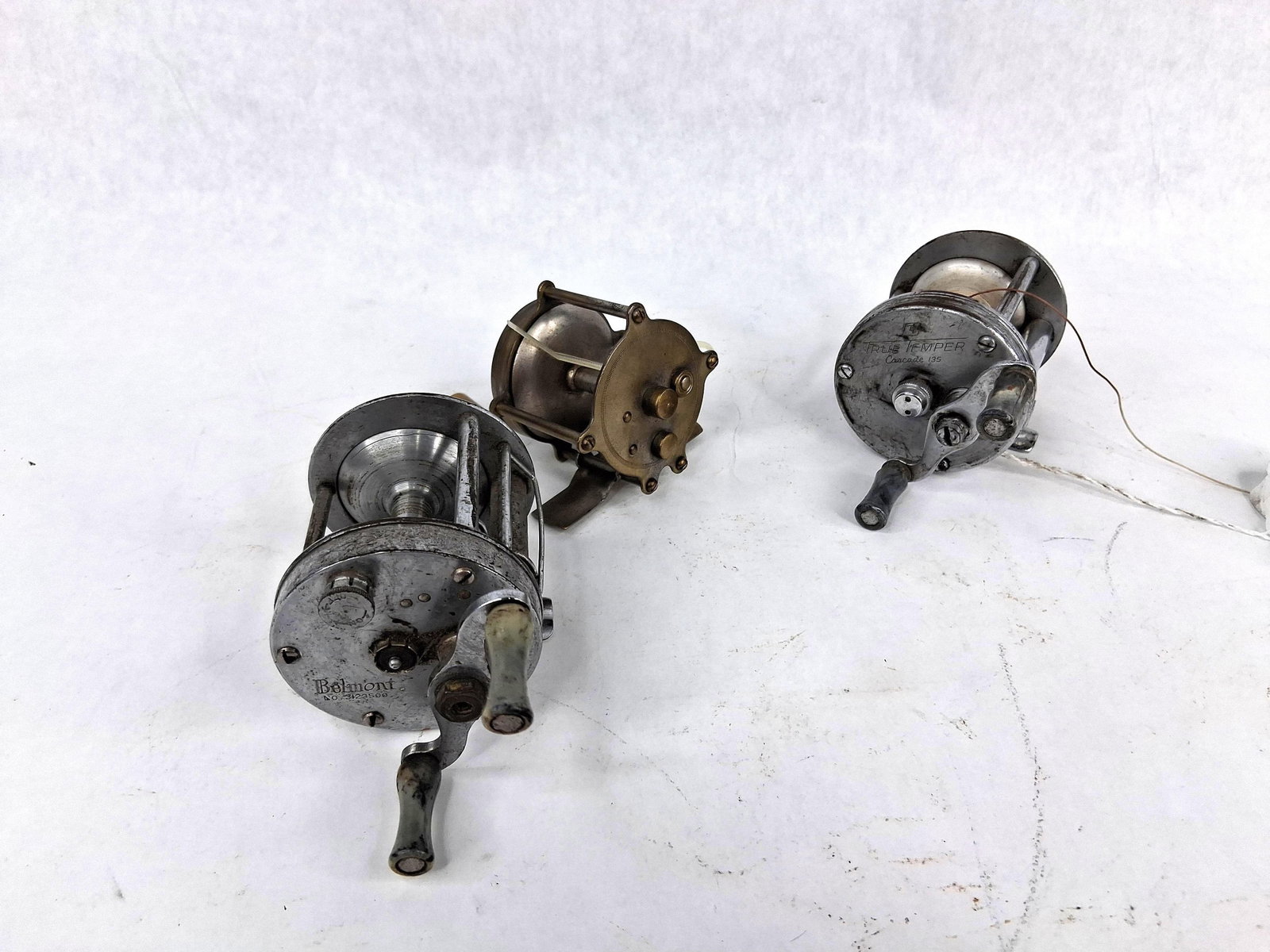 3 Vtg Reels: True Temper, Belmont, Erie: This lot comprises three baitcasting fishing reels of various makes and materials. The first reel is a silver-toned Belmont No. 3123500 model, featuring a double-handle design with marbled green and