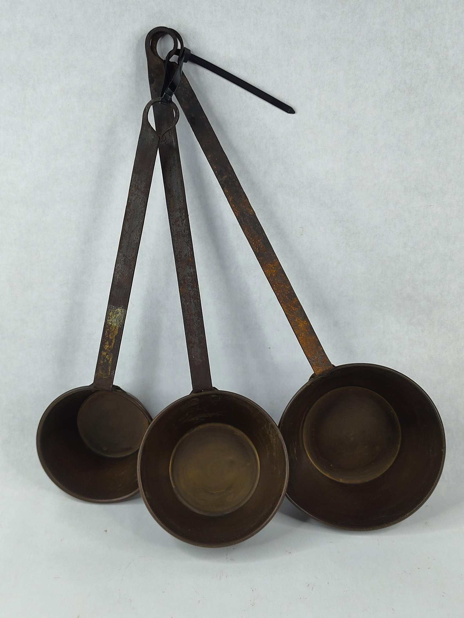 Trio Graduated Copper & Iron Ladles: A trio of graduated primitive-style dippers or measures. The circular bowls are crafted from heavy-gauge copper with a deep, aged patina and finished with rolled rims. Each bowl is attached to a long,