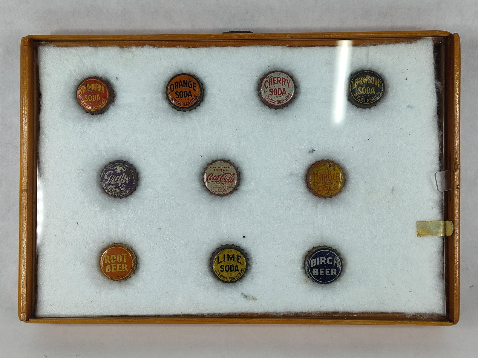 Set 10 Vtg Soda Bottle Caps Shadow Box: A collection of ten lithographed tin-plated steel bottle crown caps presented in a wooden shadow box display case. The assortment features a variety of beverage flavors and brands characteristic of