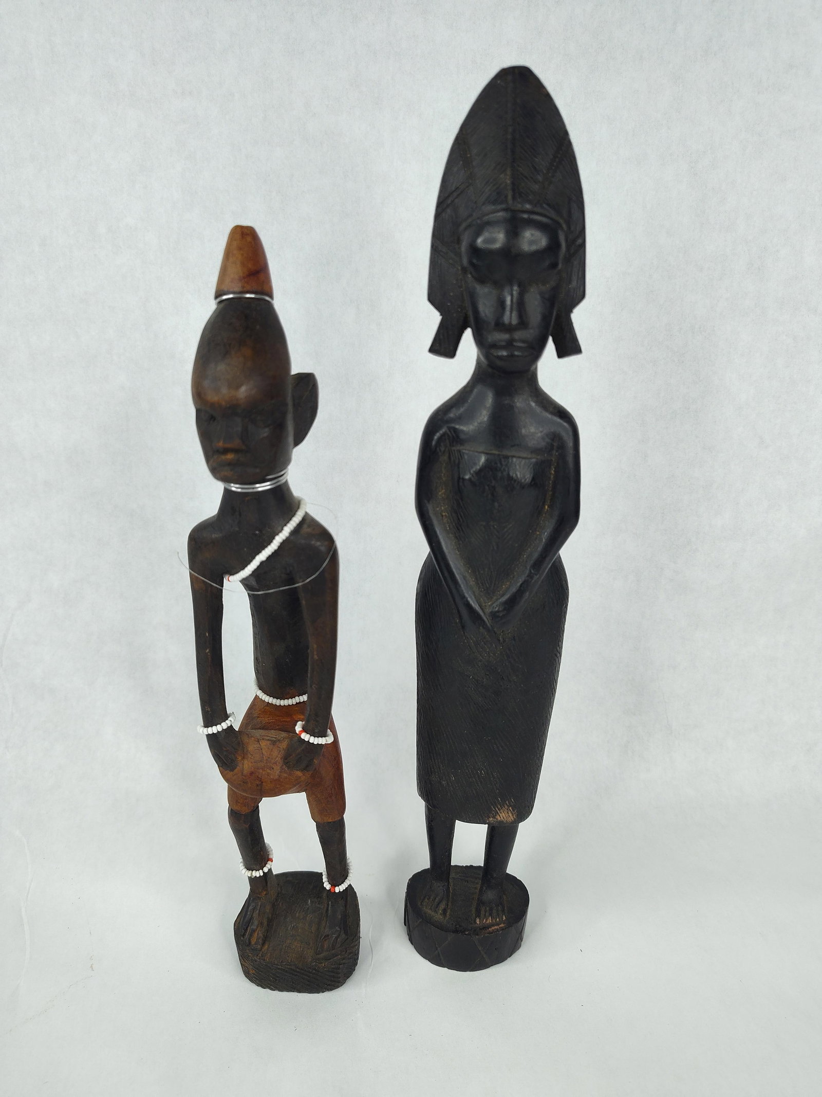 Pair African Carved Wood & Mixed Media: This lot consists of two hand-carved wooden figures of African origin. The taller female figure is fashioned from a dense, dark-toned wood with an ebonized finish. She is depicted standing with hands