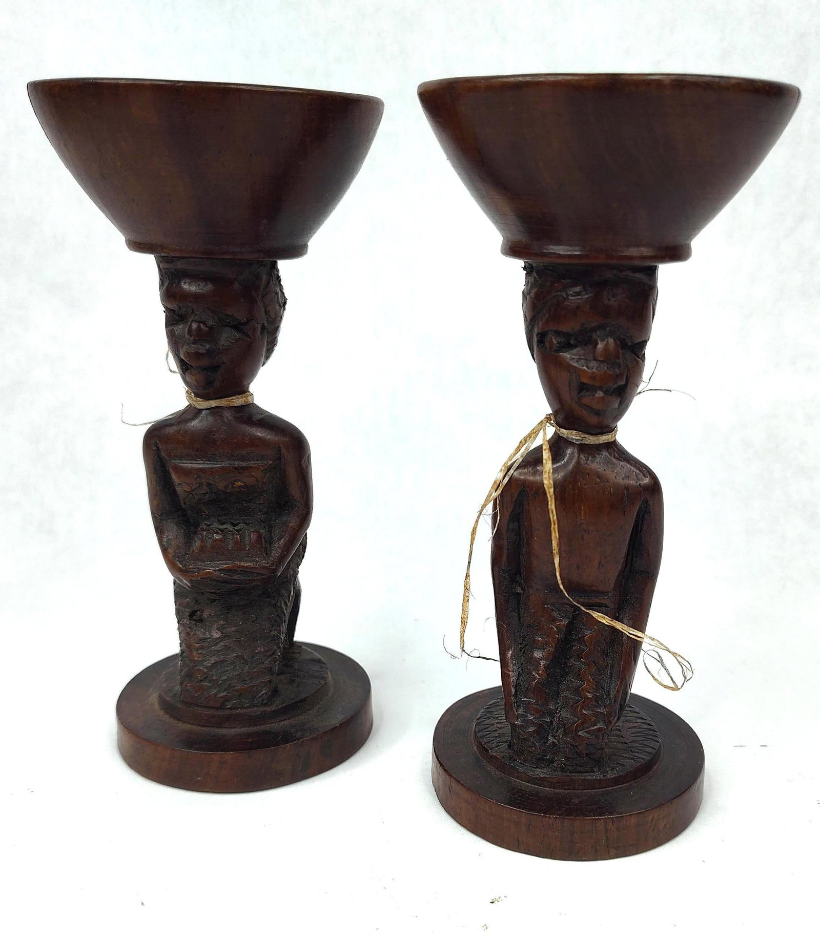 Pair African Carved Wood Candle Holders: A pair of hand-carved wooden candle holders representing a male and female figure in traditional kneeling poses. Each figure supports a flared, bowl-shaped candle socket on its head, which includes a