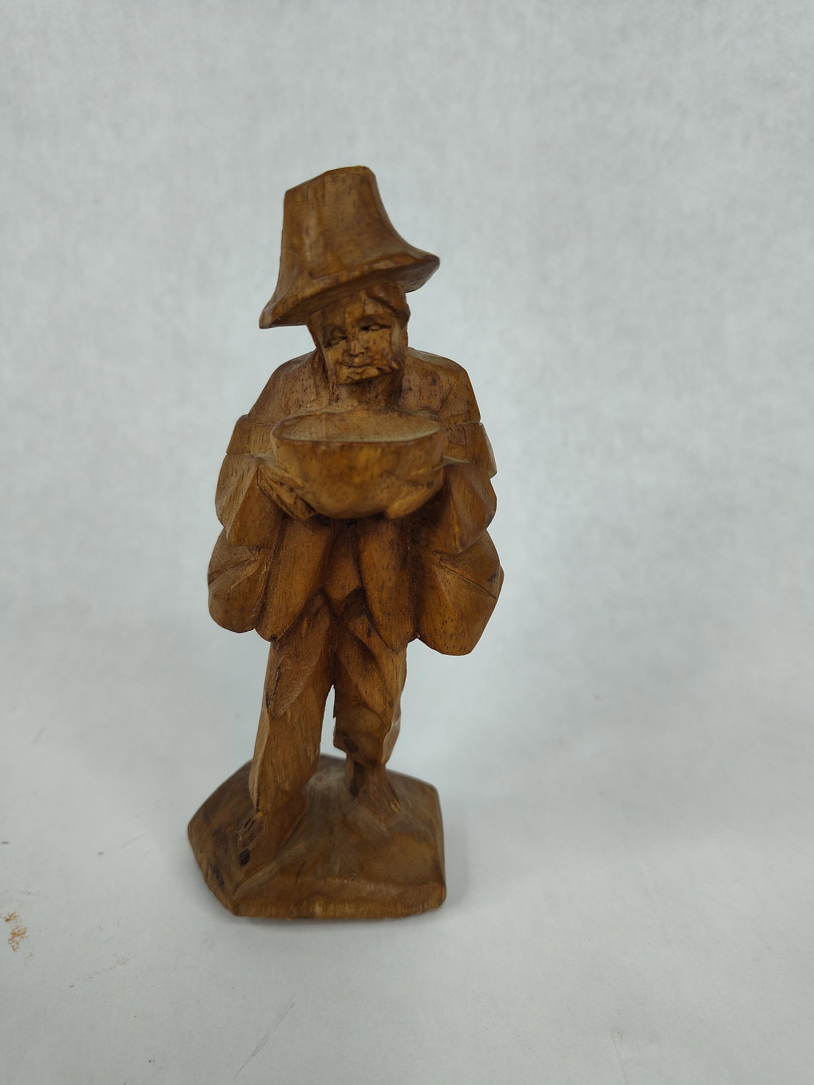Hand-Carved Wood Traveling Folk Figure: This hand-carved wooden sculpture depicts a male figure in rustic attire, featuring a tall, tapered hat, a hooded cloak, and a backpack. The figure is portrayed in a standing position, holding a wide,