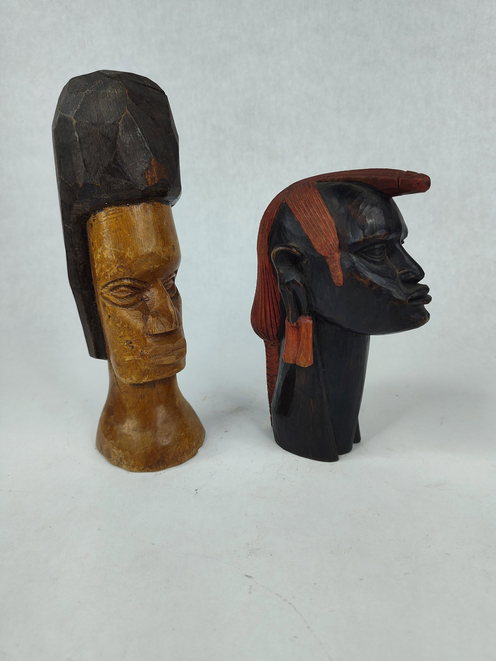 Pair Vtg Hand Carved African Wood Busts: This pair of hand carved wooden busts consists of two stylized human heads, likely produced as decorative folk art or souvenir carvings. The first bust is carved from a honey-toned wood and features a
