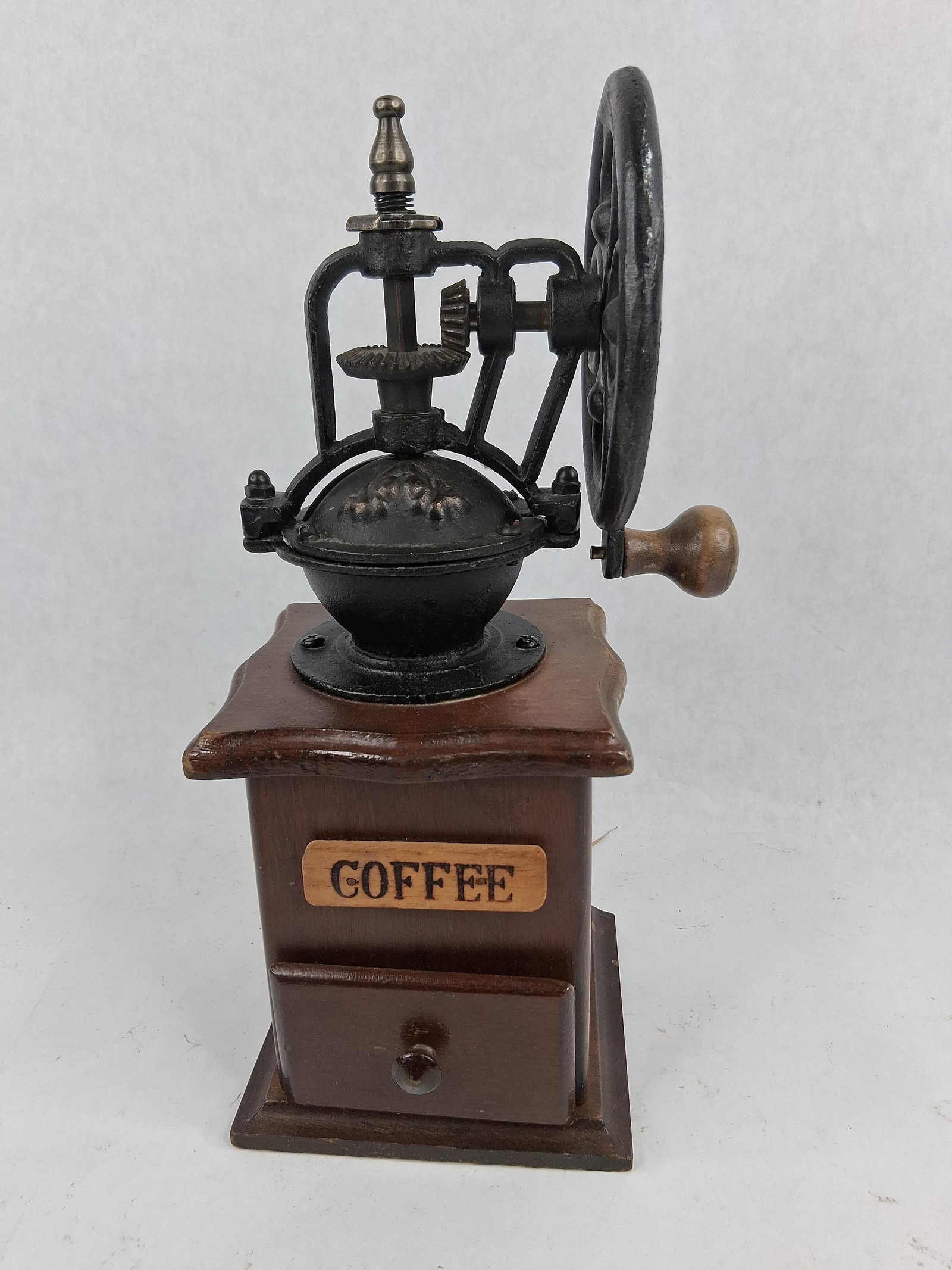 Flywheel Coffee Mill, Cast Iron & Wood: This manual flywheel coffee mill is a tabletop kitchen collectible constructed from cast iron and finished wood. The device features a decorative cast-iron flywheel with scrolling openwork and a turne