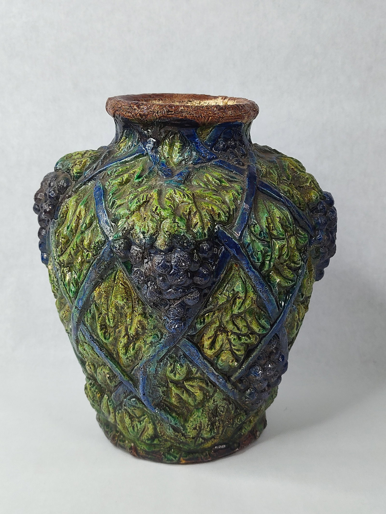 Art Pottery Majolica Grapevine Vase: A majolica art pottery vase featuring high-relief decoration. The ovoid earthenware body is decorated with a molded and hand-modeled fruiting grapevine motif. A trellis of raised deep blue glazed band