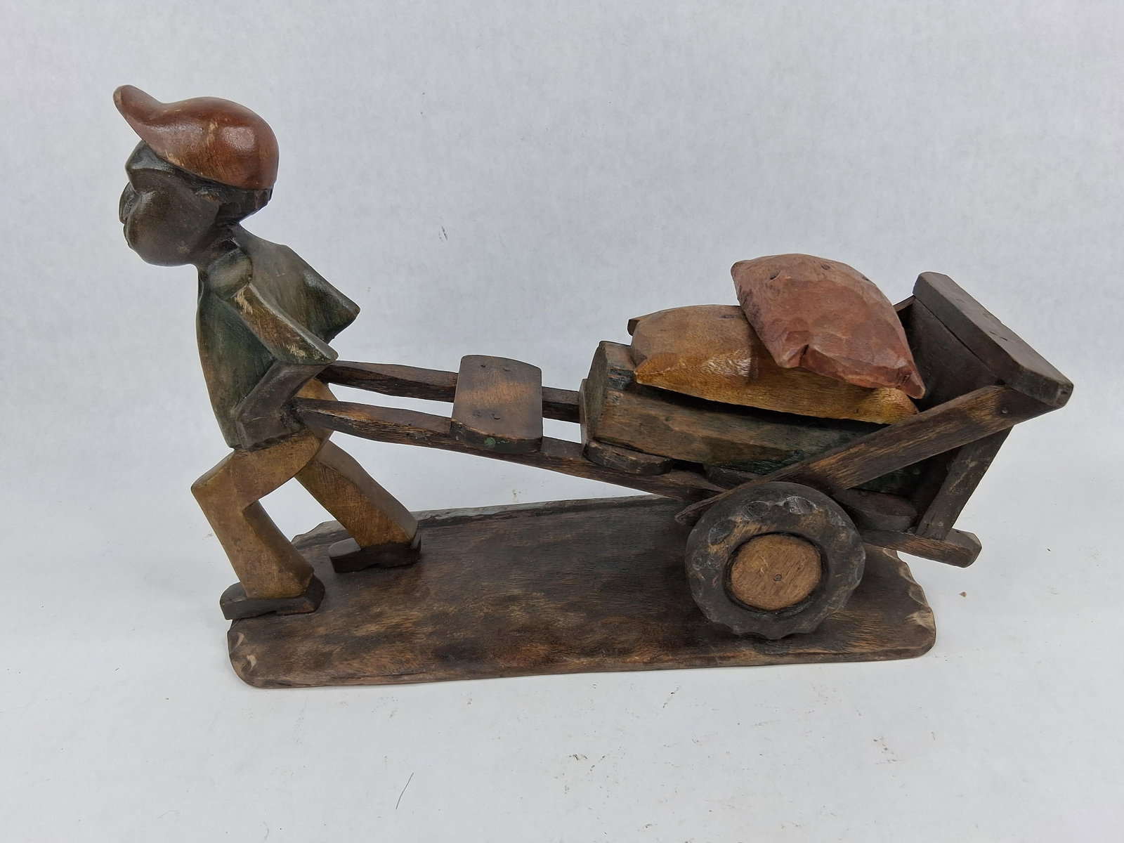 Hand Carved Wood Man w/ Wagon Sculpture: This hand carved wood folk art sculpture depicts a stylized male figure of African descent pulling a two-wheeled cart laden with three distinct cargo blocks representing sacks or stones. The sculpture