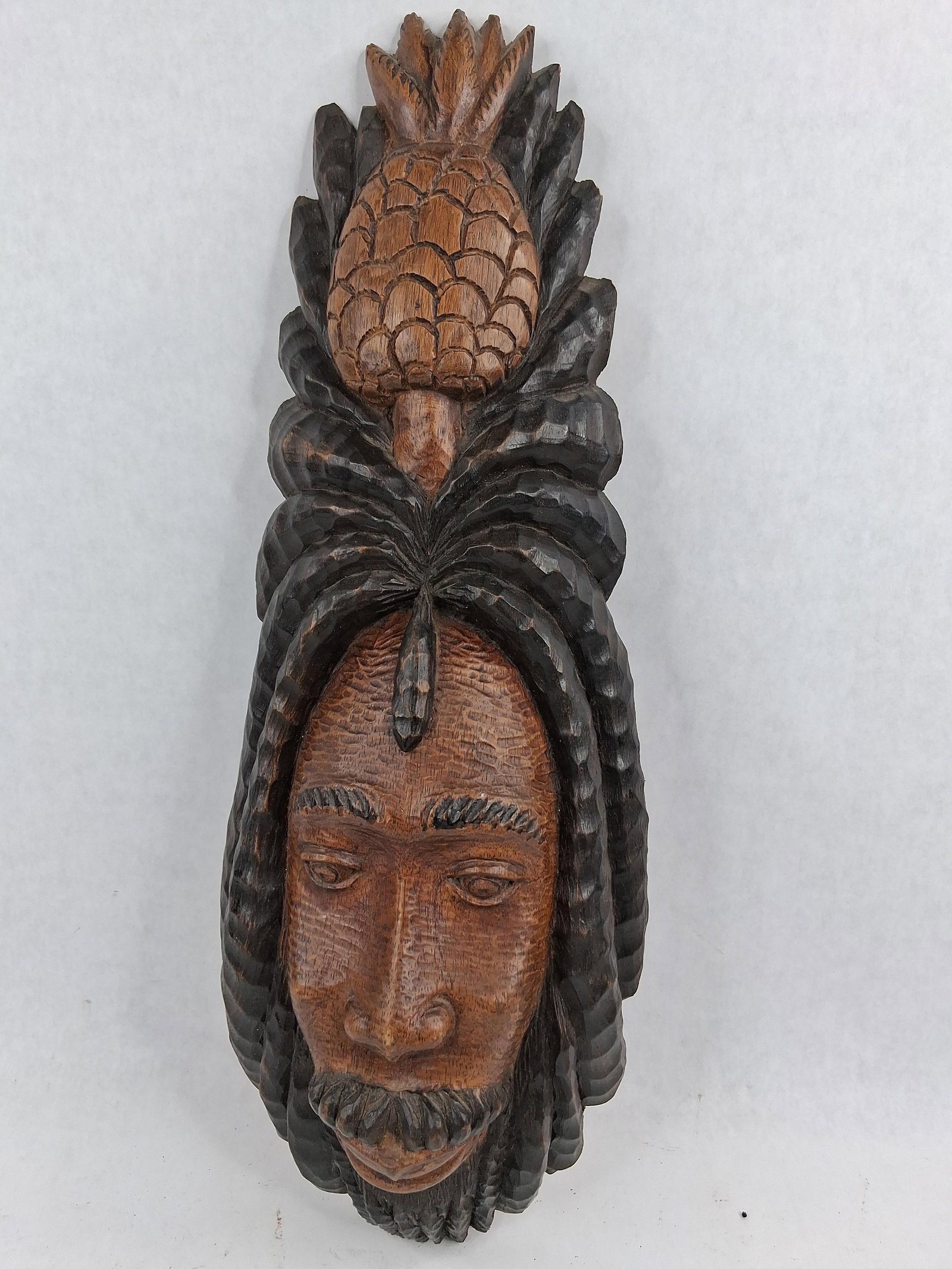 Jamaican Folk Art Rasta Wood Plaque: This hand-carved wooden wall plaque is a work of Jamaican folk art, featuring a stylized portrait of a man with dreadlocks surmounted by a large pineapple. The piece is constructed from wood and