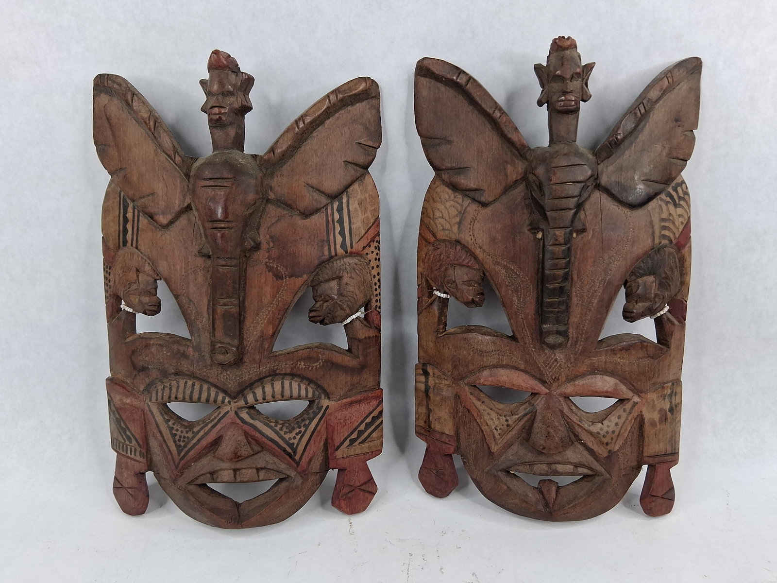 Pair African Style Elephant/Human Masks: A pair of hand-carved wooden decorative masks featuring a complex vertical arrangement of zoomorphic and anthropomorphic elements. Each mask is topped with a small human head supported by the long