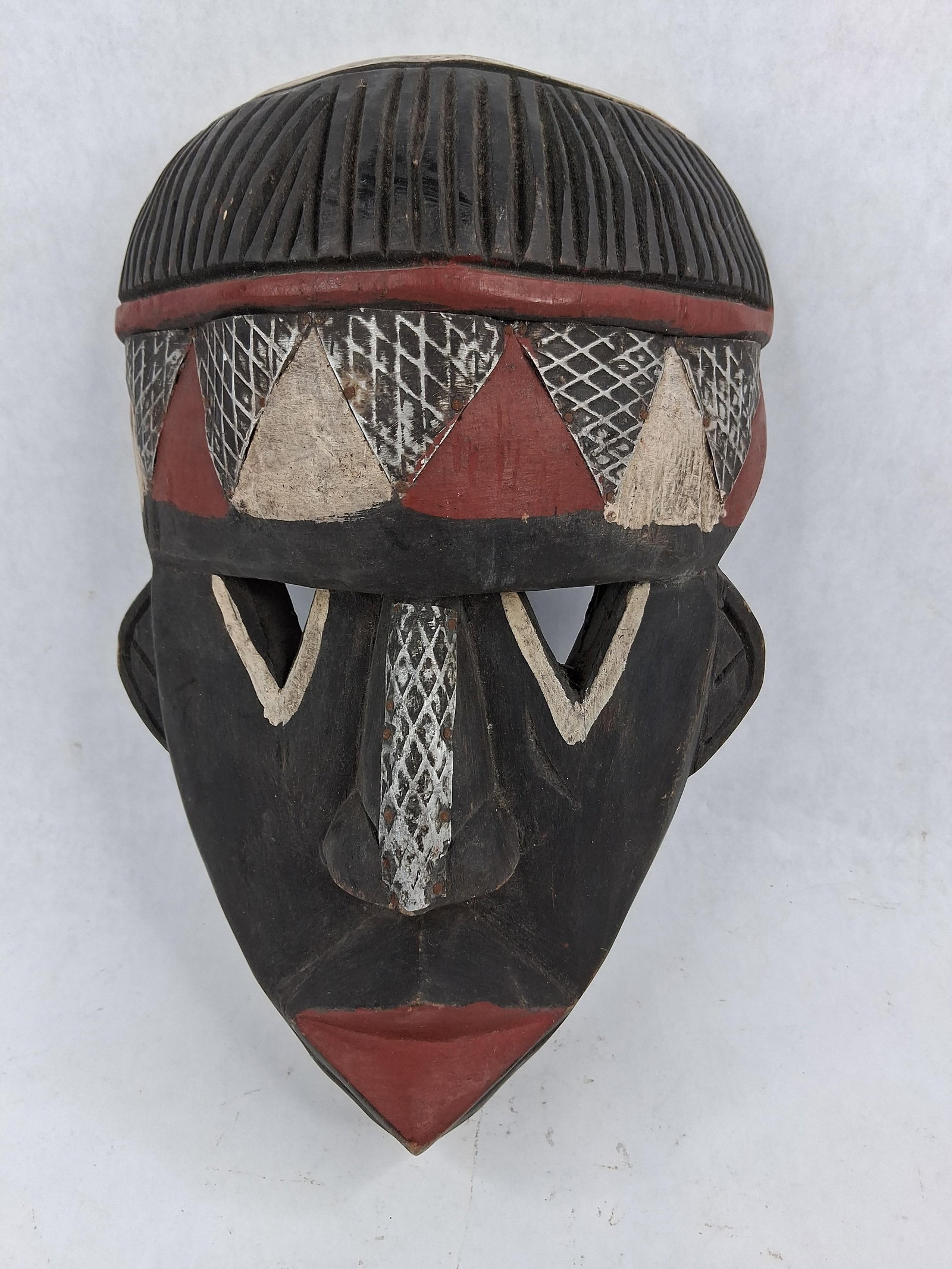 Ghanaian Carved Wood Geometric Mask: This is a hand-carved wooden decorative mask originating from Ghana, as indicated by a label on the reverse. The mask is carved from a single block of wood with a dark-stained finish and features bold
