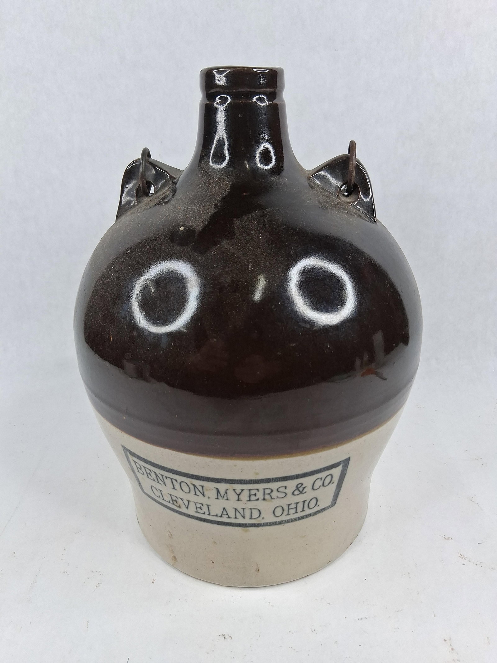 Benton, Myers & Co. Advertising Jug: A two-tone stoneware shoulder jug featuring a globular body and a short, narrow neck with a flared, double-ringed rim. The vessel is finished with a dark brown Albany-type slip glaze on the upper