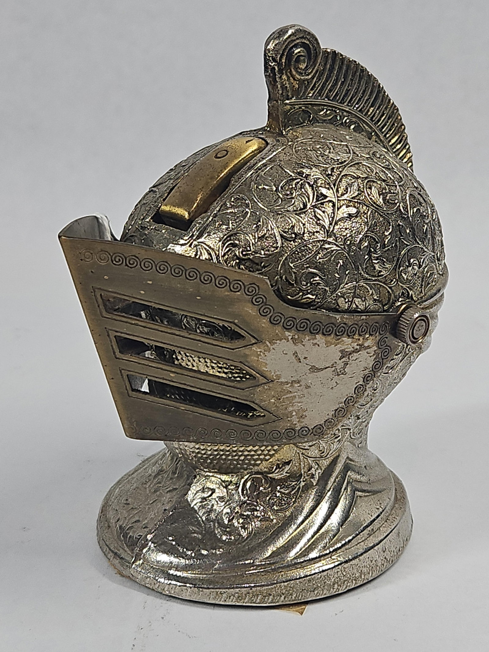 Japanese Metal Knight Helmet Lighter: A Japanese cast metal novelty table lighter fashioned in the form of a knight's close helmet with a decorative comb plume. The object is constructed from a zinc-based alloy with a two-tone plated