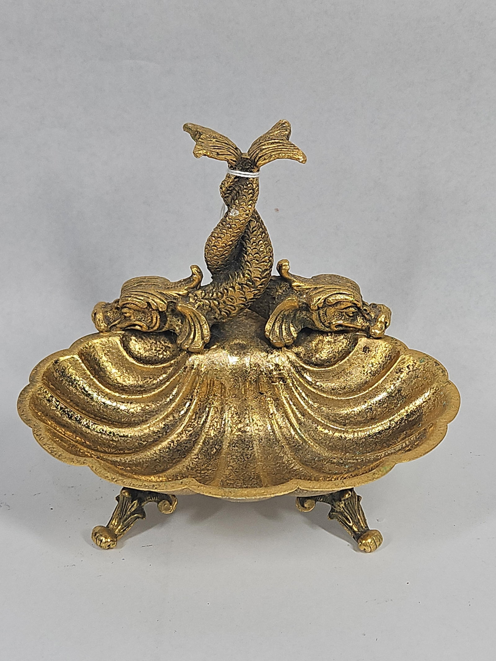 Cast Brass Double Shell Dolphin Dish: A cast brass decorative dish featuring two adjoined scallop-shaped basins joined at a central point. The piece is surmounted by a prominent figural handle depicting two stylized mythological dolphins