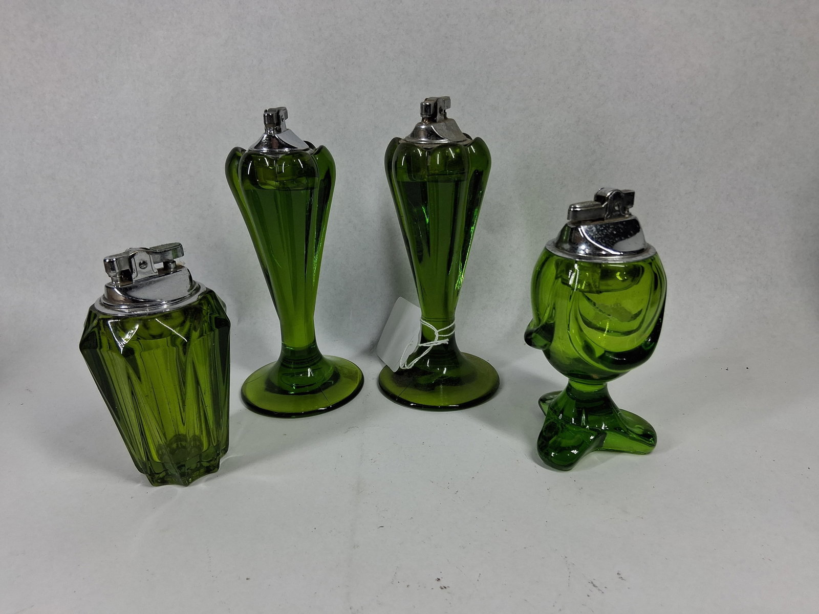 Set 4 Mid-C. Green Glass Table Lighters: An assortment of mid-century modern pressed glass table lighters in a vibrant emerald green color. The group features three distinct forms: one squat lighter with vertical tapering facets and a hexago