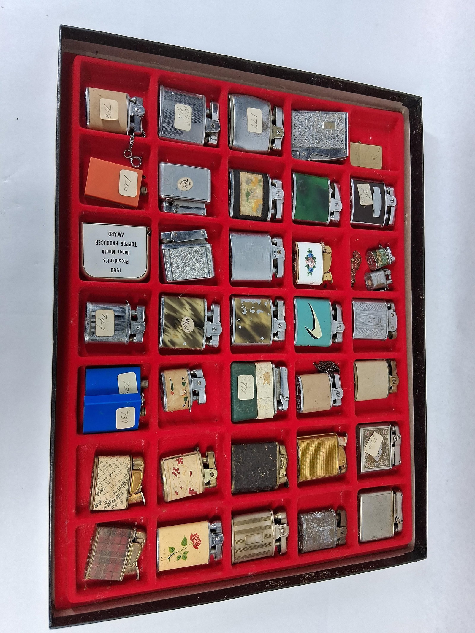 Ronson, Colibri & Champion Vtg Lighters: An assortment of vintage pocket lighters presented in a red felt-lined display case. The collection encompasses various mechanical designs including automatic push-button and lift-arm systems.