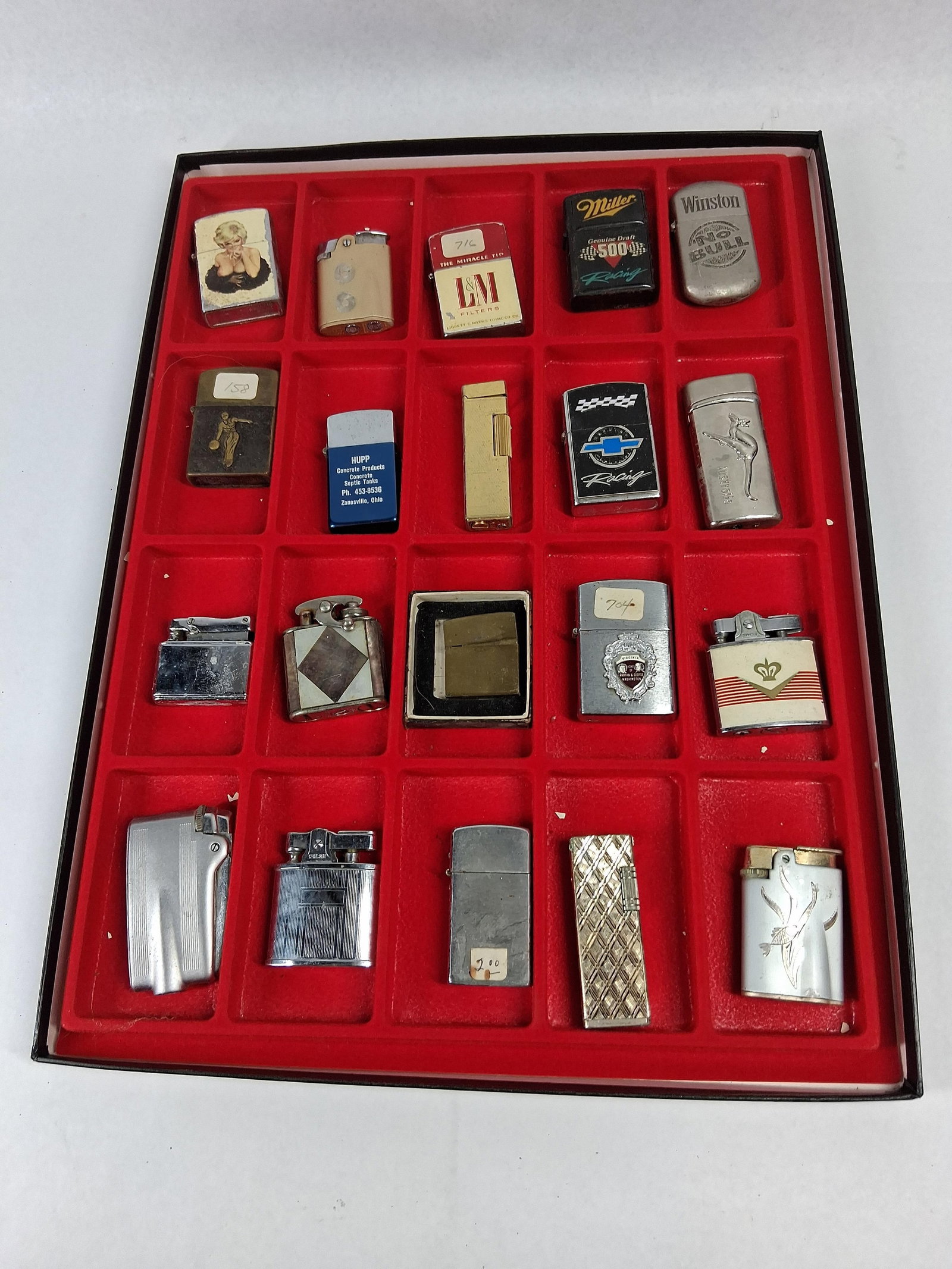 20 Vtg Ronson Penguin Trilite Lighters: A collection of twenty vintage pocket lighters presented in a red flocked display tray, primarily constructed from chrome-plated steel, brass, and aluminum with various finishes including enamel,