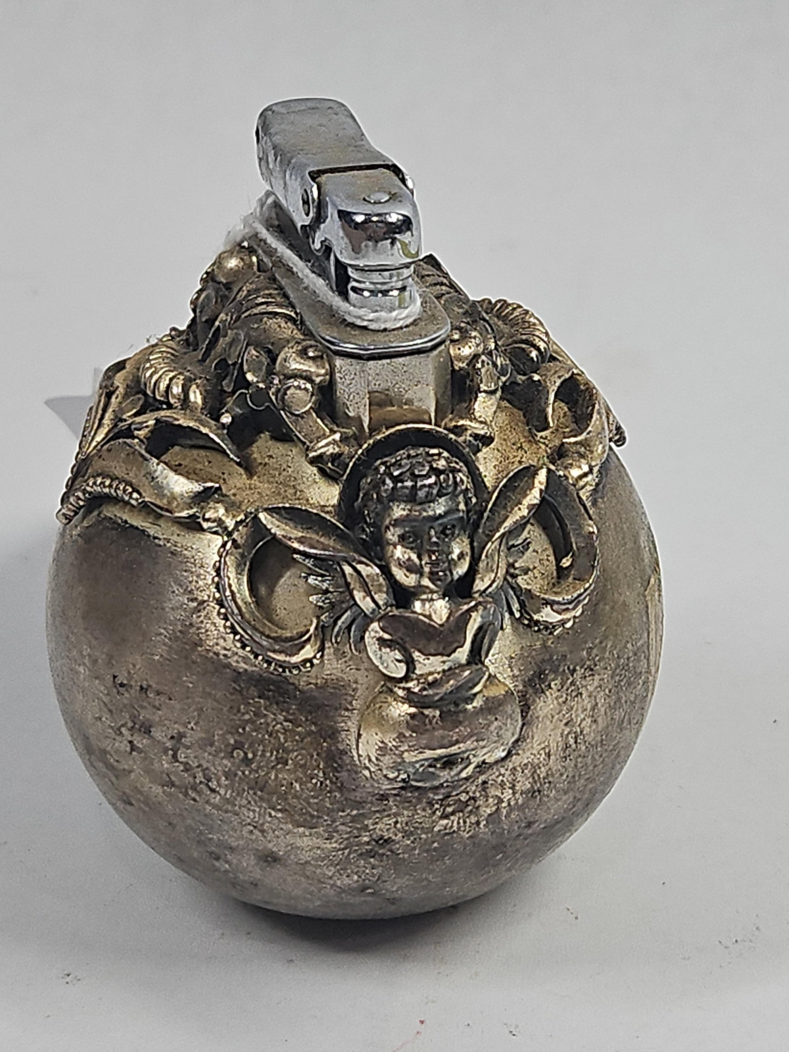 Vtg Silver Toned Cherub Table Lighter: This vintage spherical table lighter features a cast metal base with a silver toned finish, heavily ornamented with high relief cherub figures and neoclassical scrollwork. The lighter consists of a we