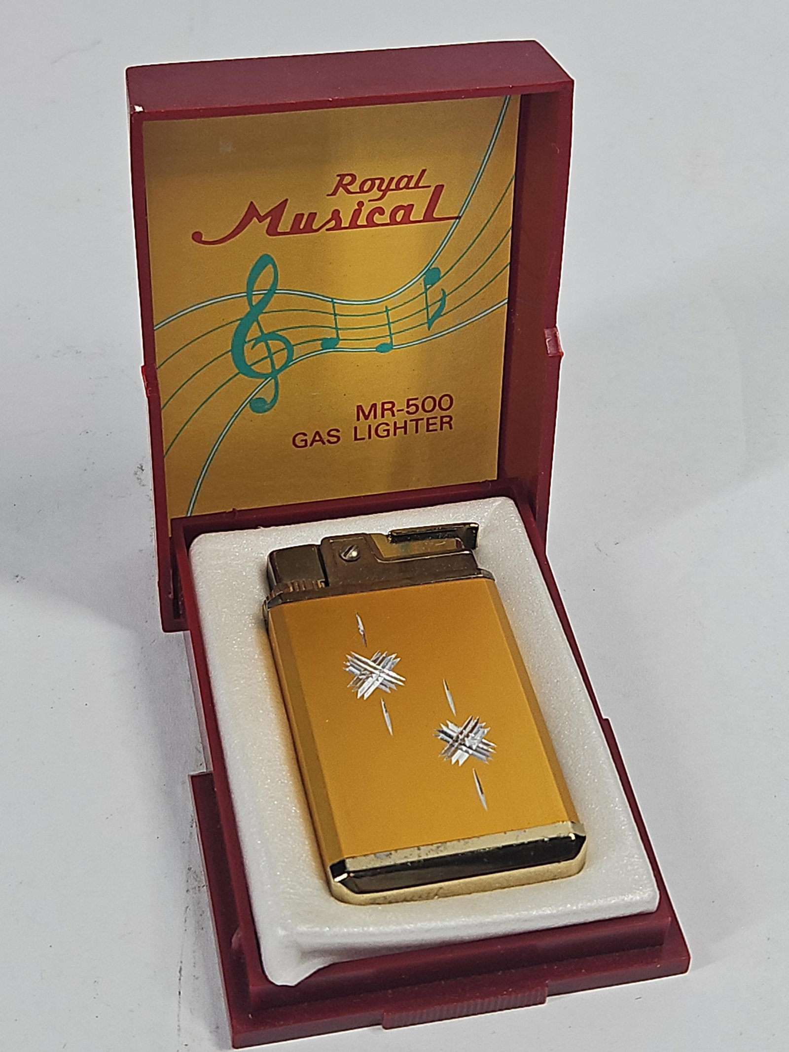 Royal Musical MR-500 Music Box Lighter: A Japanese gas lighter with an integrated music box mechanism, model MR-500. The lighter is constructed with a rectangular gold-toned metal body featuring a brushed finish and decorated with two