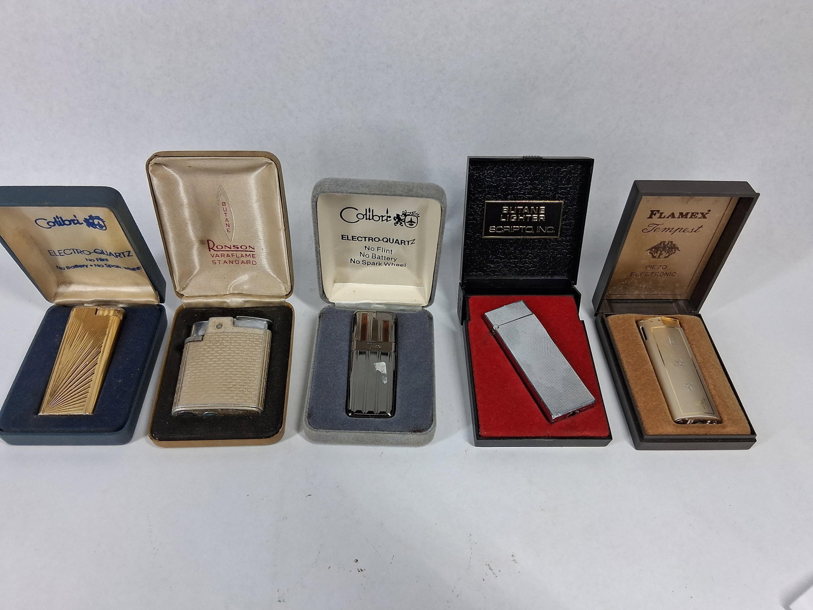 Vtg Ronson, Colibri & Scripto Lighters: A collection of vintage pocket lighters featuring various ignition mechanisms, each presented in a manufacturer-branded box. The set includes a gold-tone lighter with a radial sunburst pattern; the