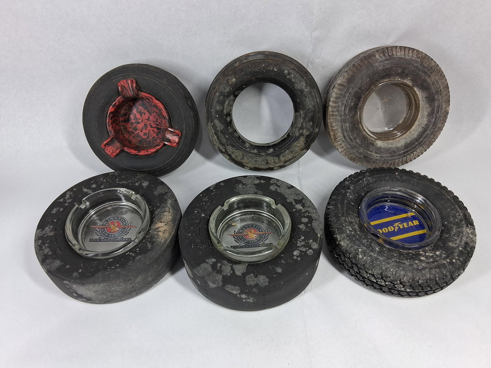 6 Vtg Goodyear/Firestone Tire Ashtrays: This lot consists of six vintage automotive-themed tire ashtrays composed of rubber, glass, and plastic. The collection includes two racing slick style tires embossed with SLICKS and NO. 1 on the